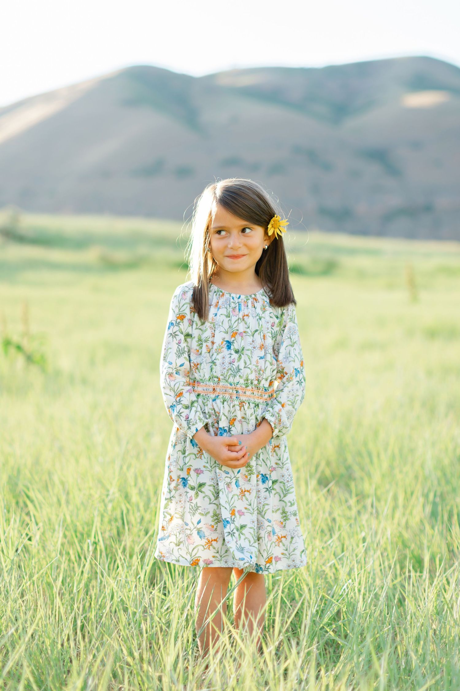 A child in a floral dress stands in a sunlit meadow with mountains in the background.