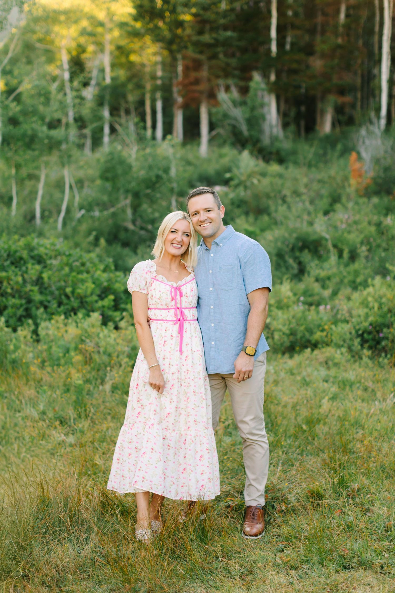 A smiling couple embraces outdoors, woman in floral dress leaning on man in blue shirt, with lush green trees behind in American Fork, Utah.