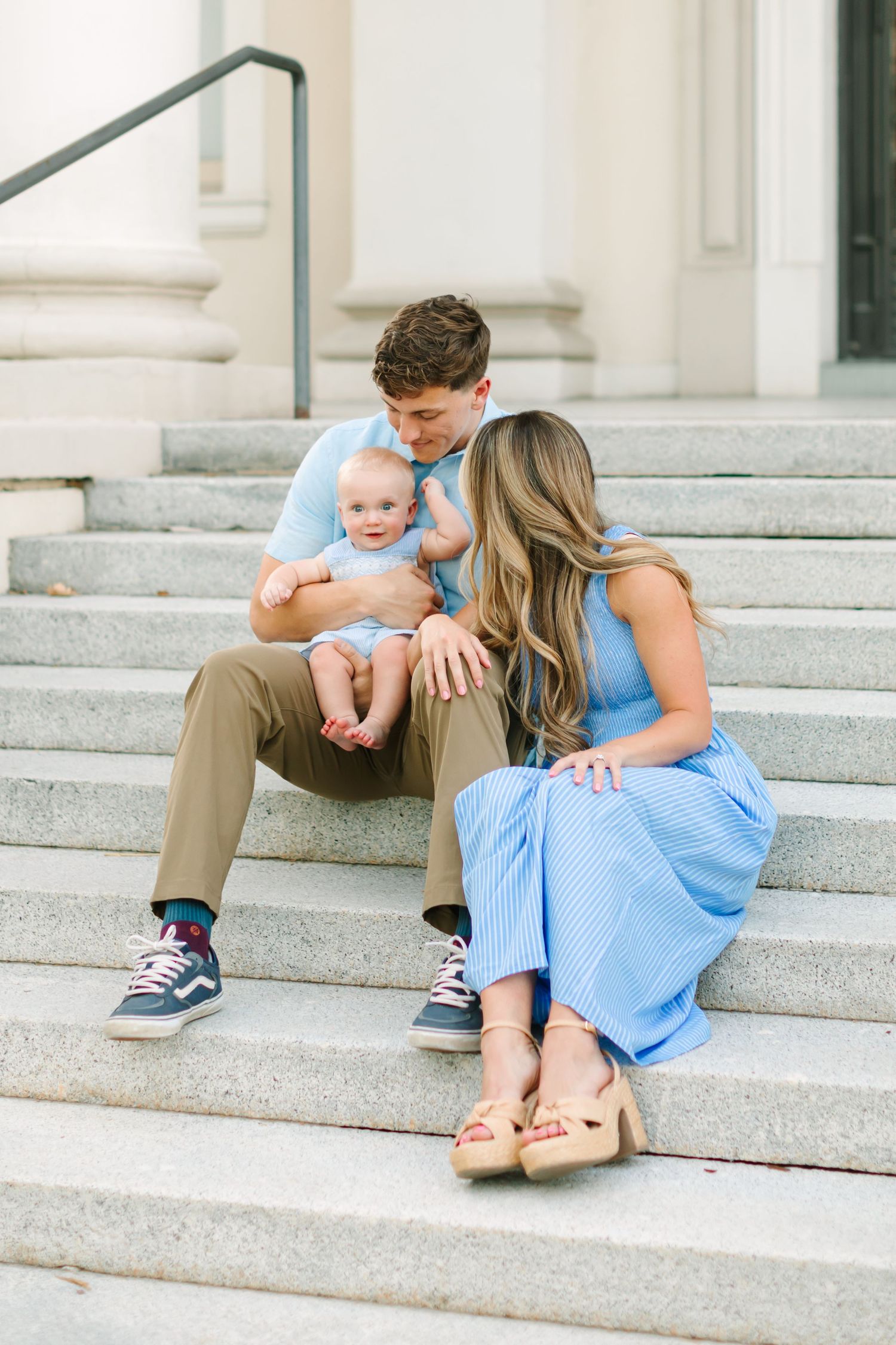 Young family with baby sitting on stone steps, parents in blue outfits sharing a tender moment with their infant.
