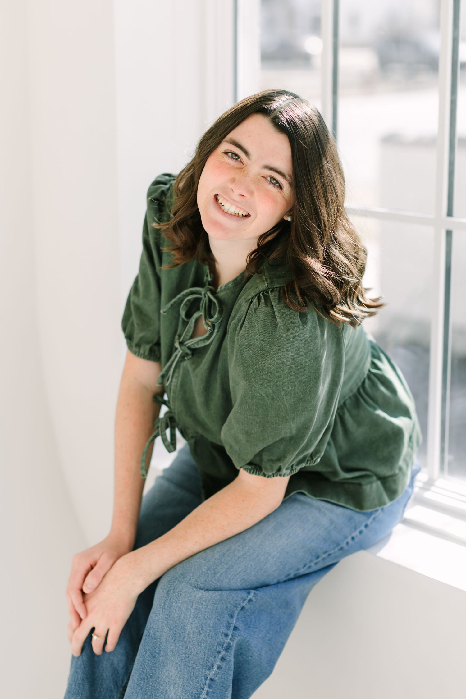 A person in a green top and jeans sits casually by a window in a bright indoor setting.