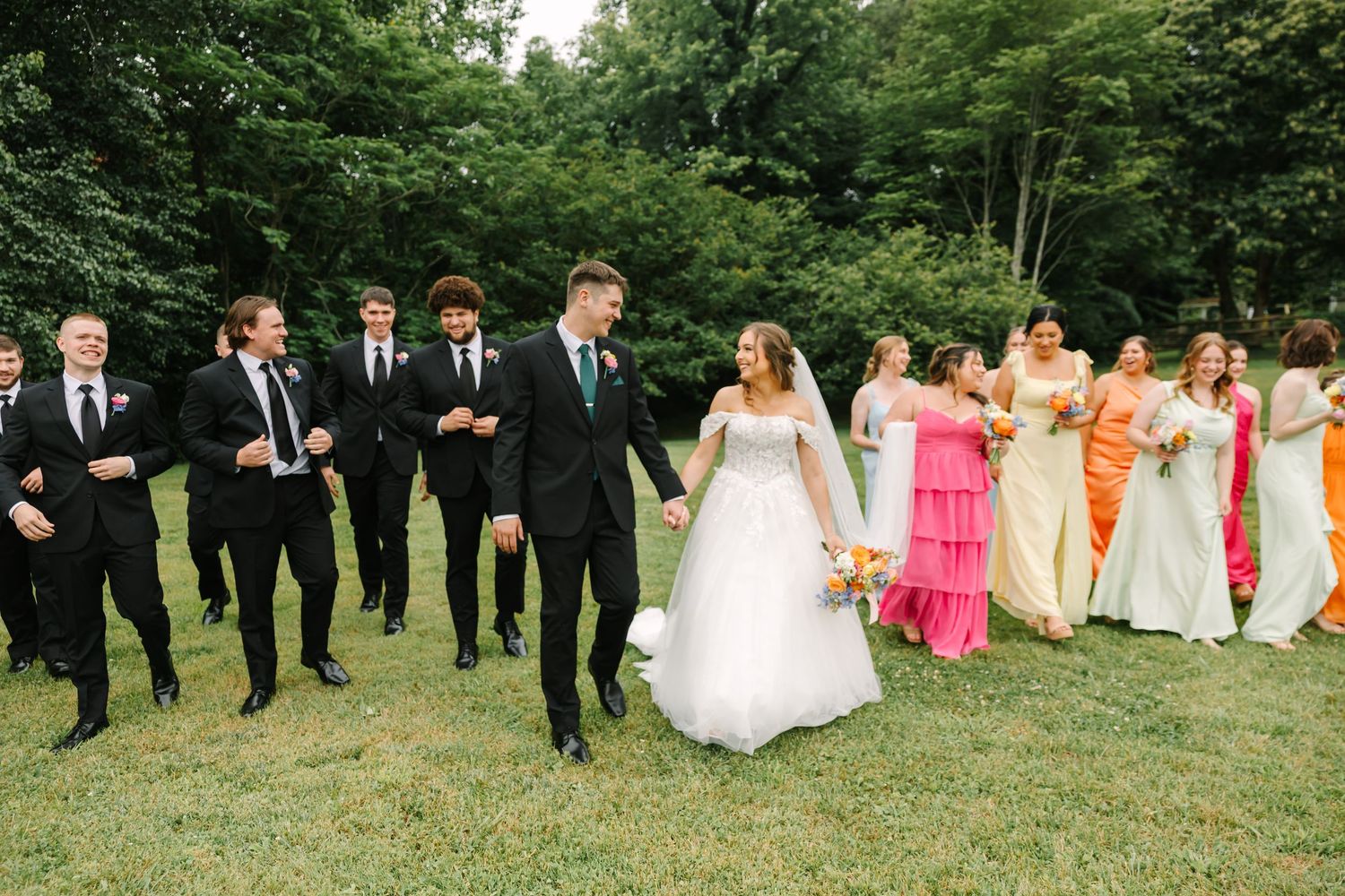 Wedding party in formal attire poses in a line across a grassy outdoor setting during a summer celebration.
