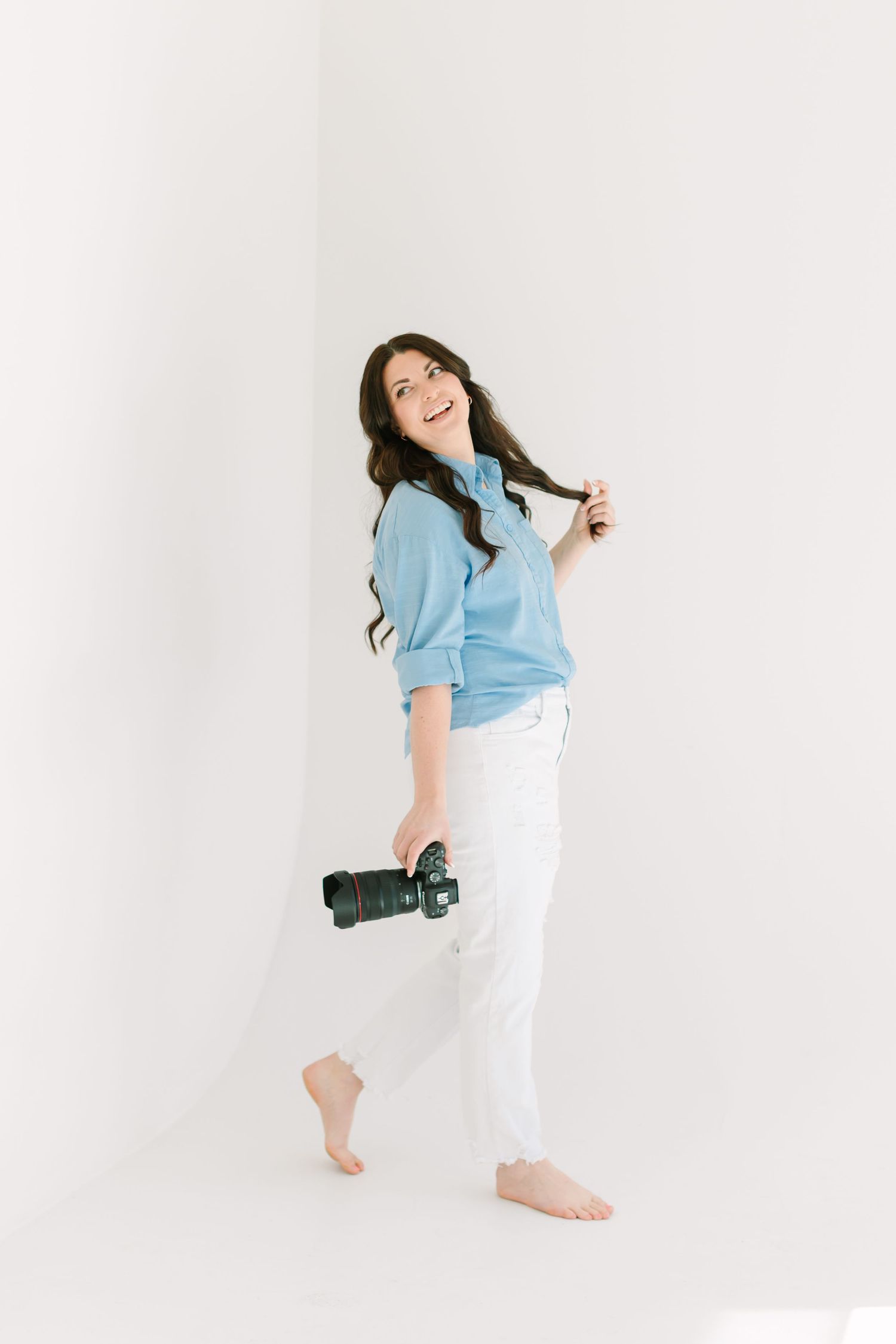 Woman in light blue sweater and white pants poses playfully against white background while holding camera.