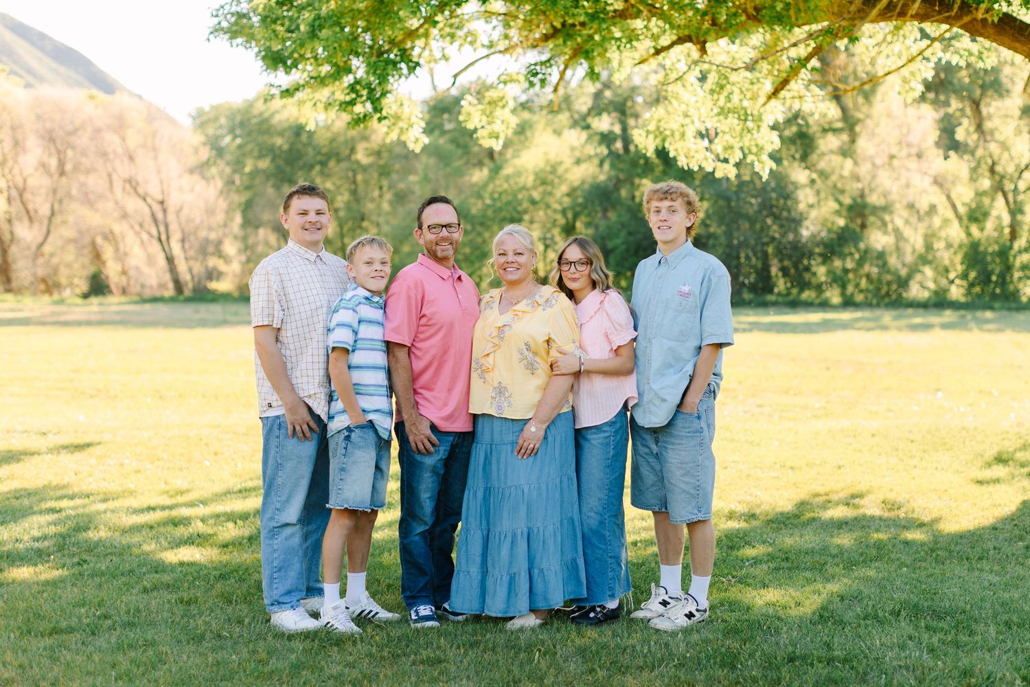 A group of people in coordinated casual outfits pose together on a grassy lawn during a sunny outdoor photo session.