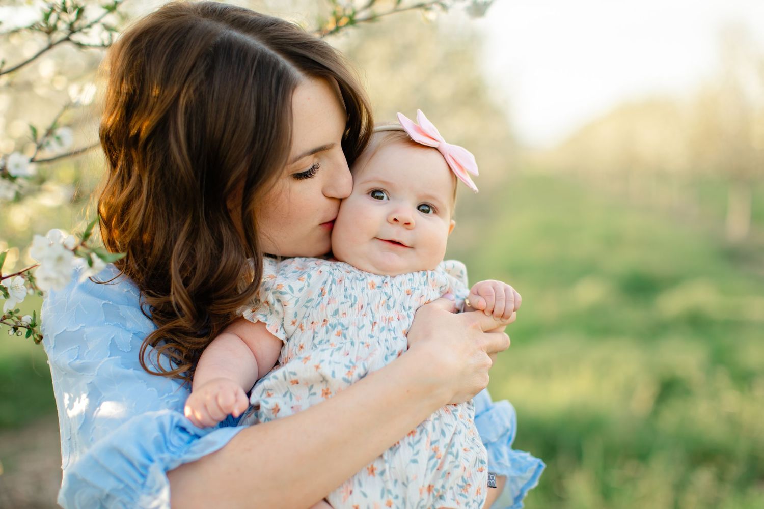 A tender moment captured outdoors as a parent embraces their baby in a floral dress during a sunny afternoon.