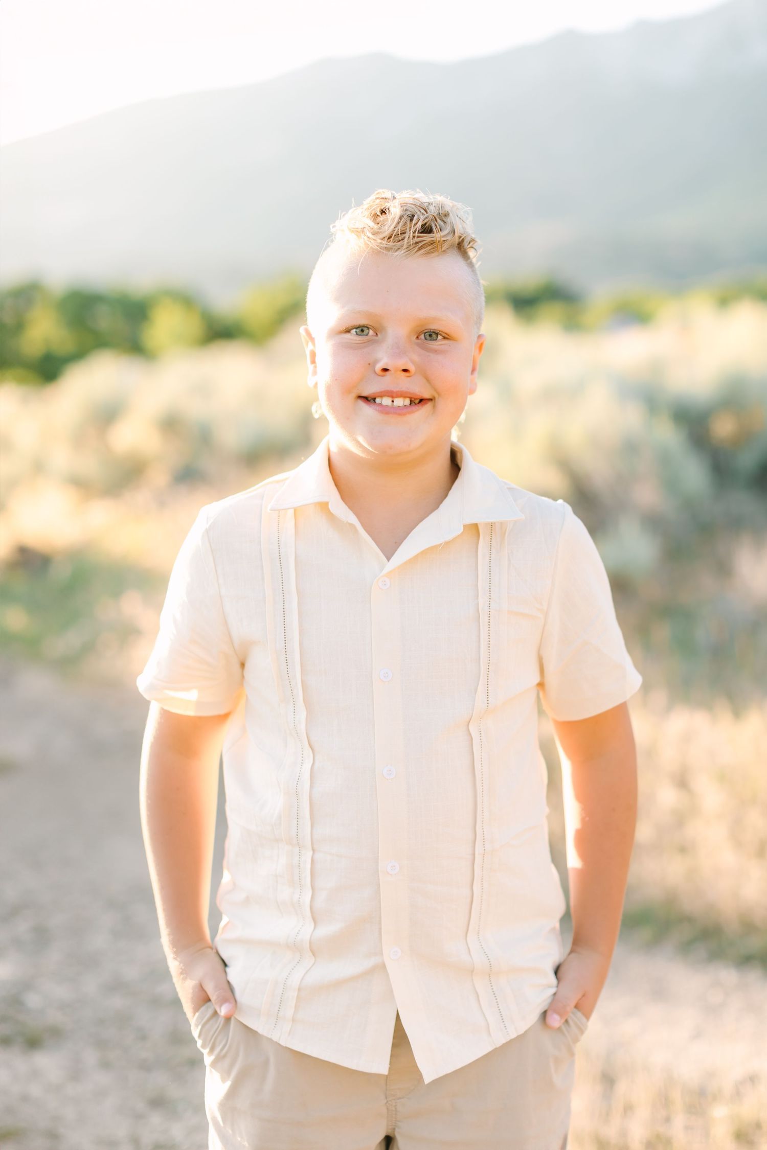 Young child in a white button-down shirt and khaki pants stands outdoors in golden sunlight with mountains in background.