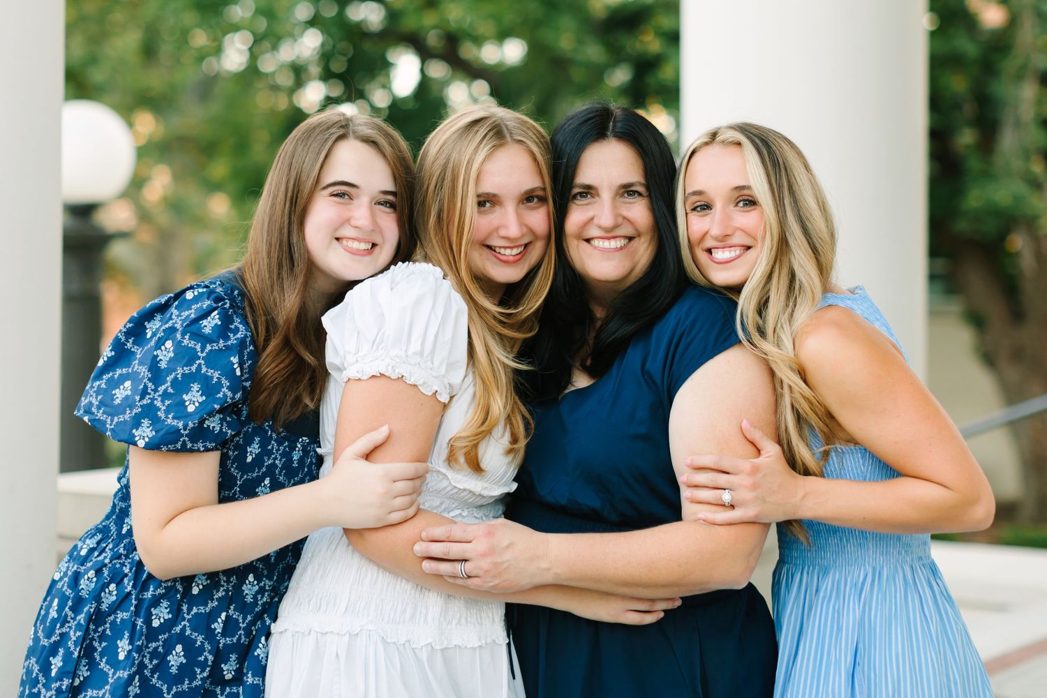 Four smiling women in blue and white dresses pose together outdoors near white columns for a family portrait.
