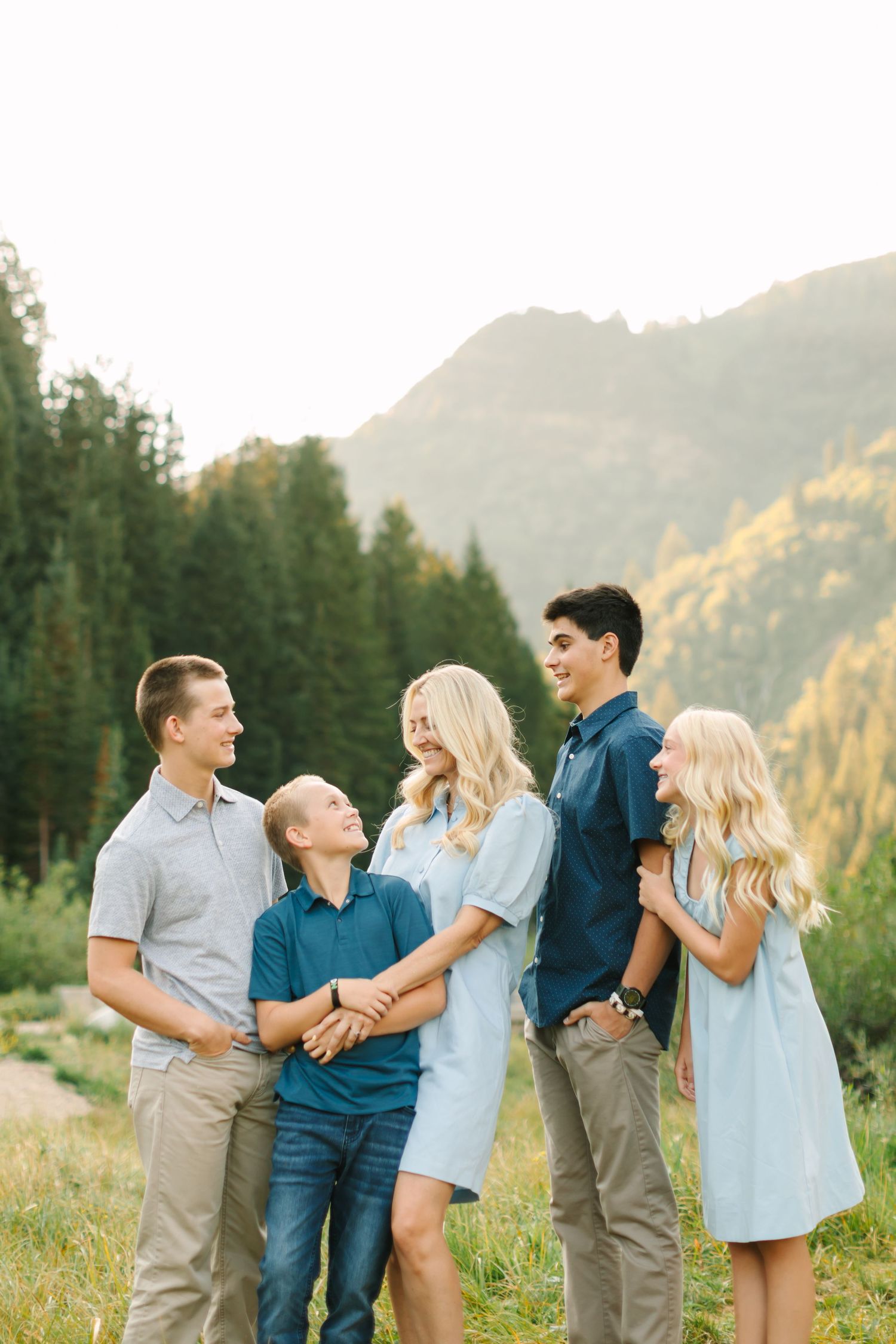 Family of five posing outdoors in mountain meadow, wearing coordinated blue outfits, smiling together with forest backdrop.