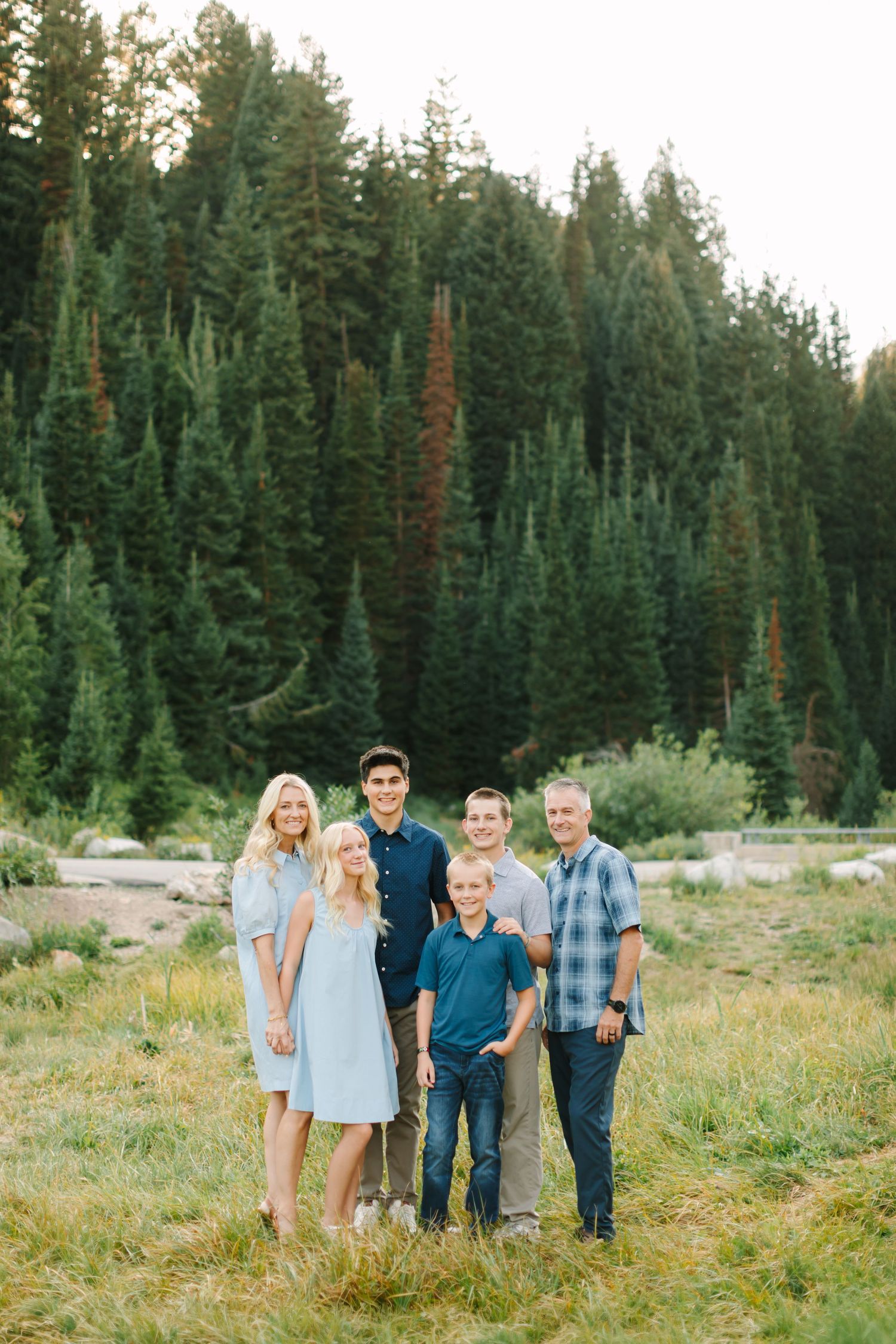 Family of six posing outdoors in a meadow surrounded by tall pine trees during golden hour in Salt Lake, Utah.