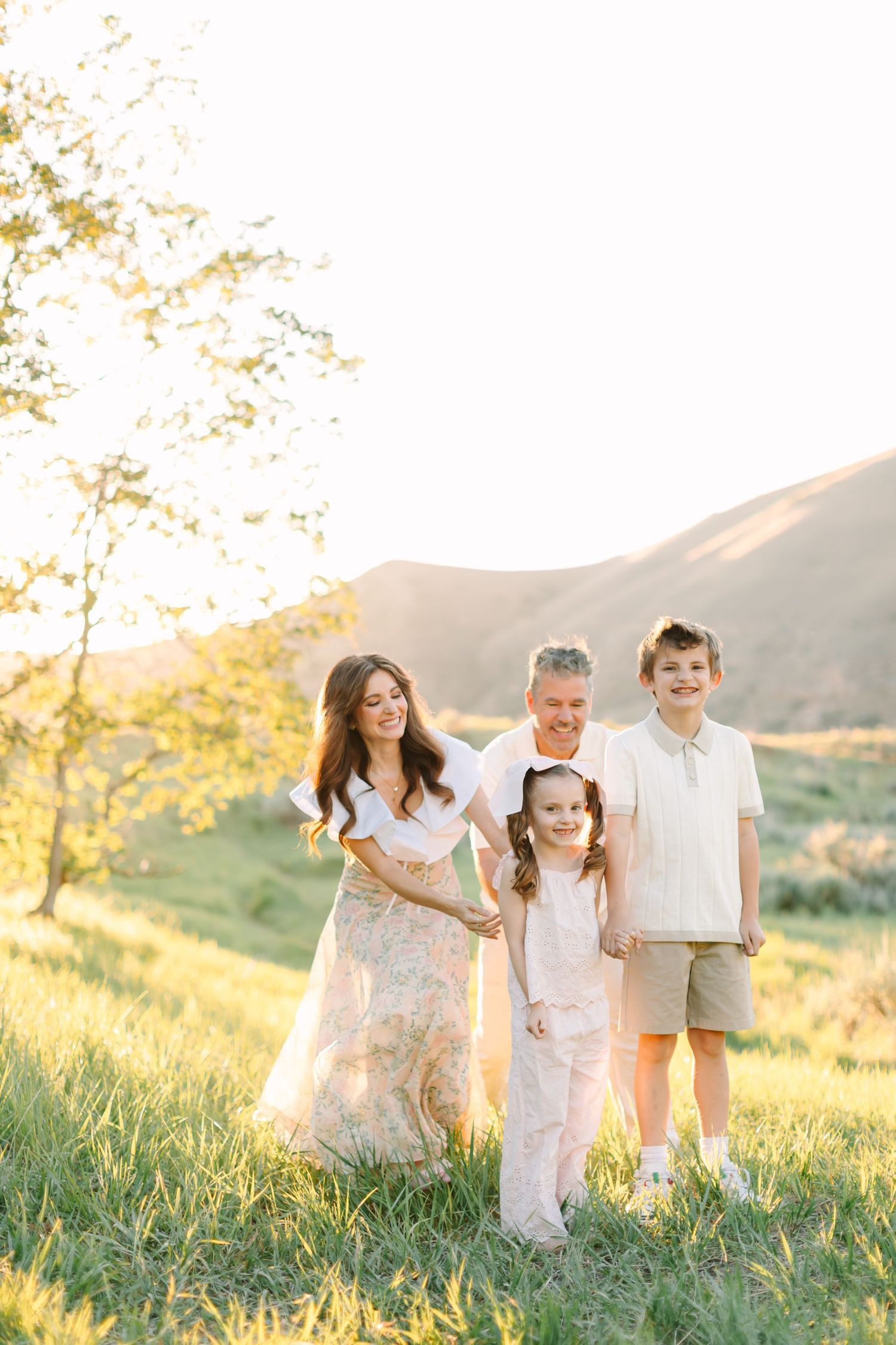 Family of four smiling outdoors in golden hour light, with green hills and trees in the background.