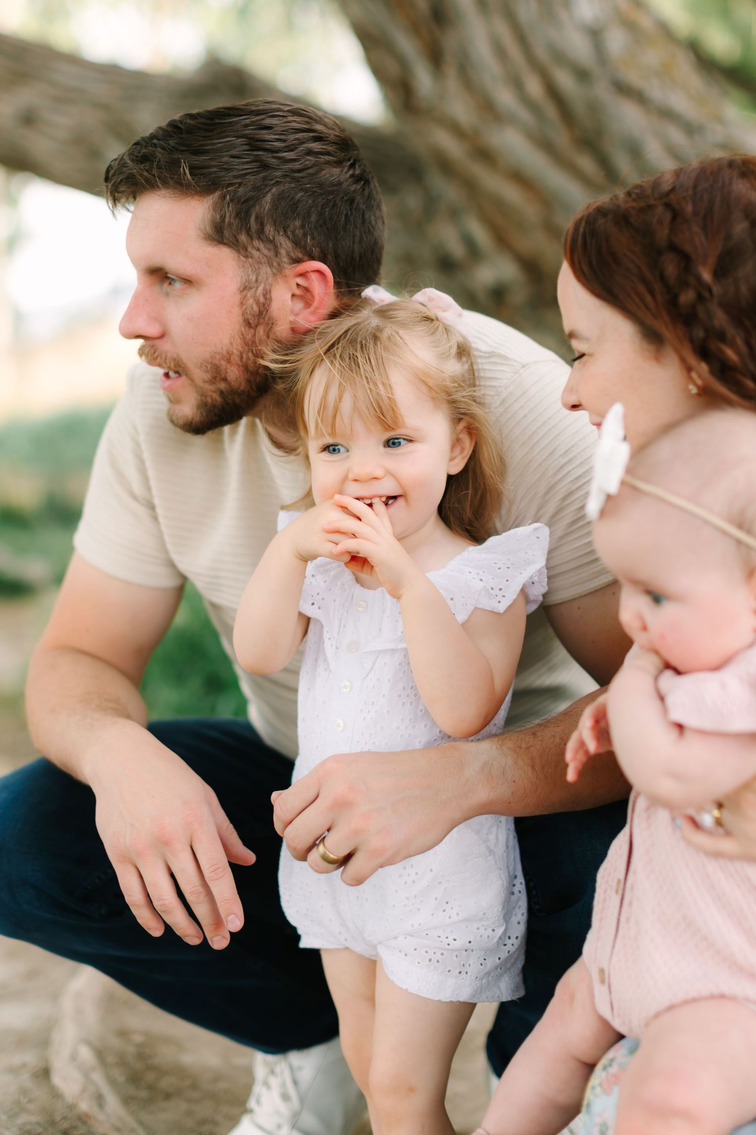 A young family with two children poses outdoors under a tree, with a toddler girl smiling at the camera.