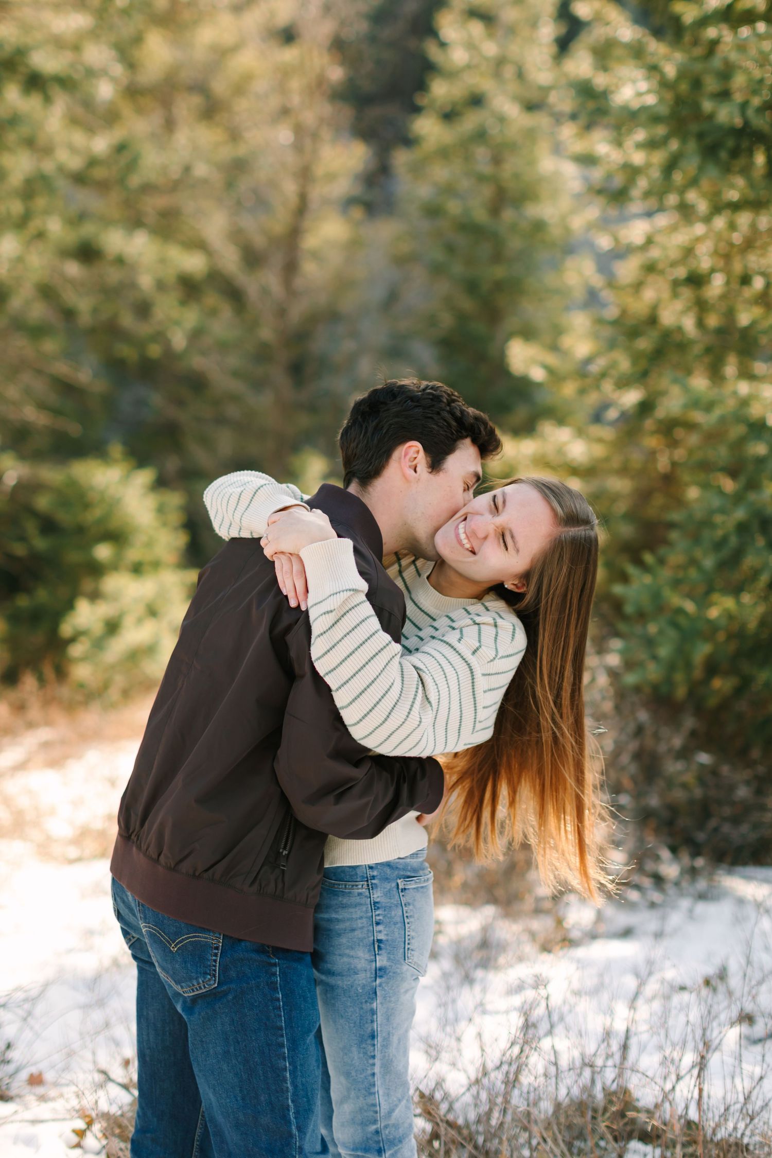 A couple embraces and laughs together on a snowy winter day surrounded by trees and sunlit foliage.