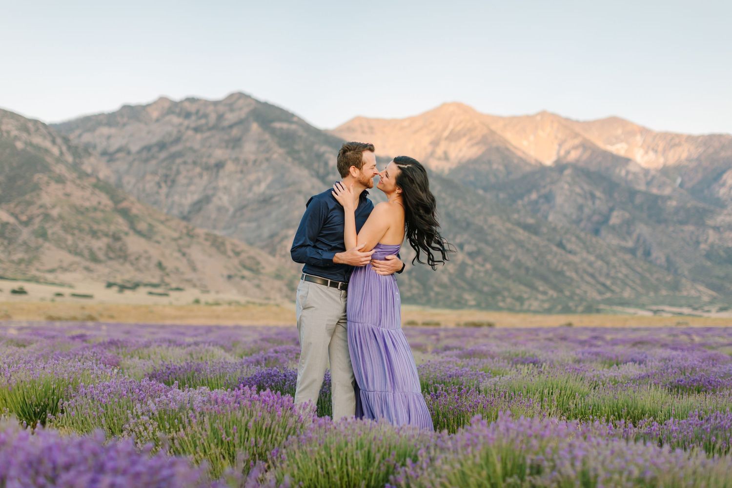 Couple in romantic poses surrounded by blooming purple lavender fields with mountain landscape in the background.