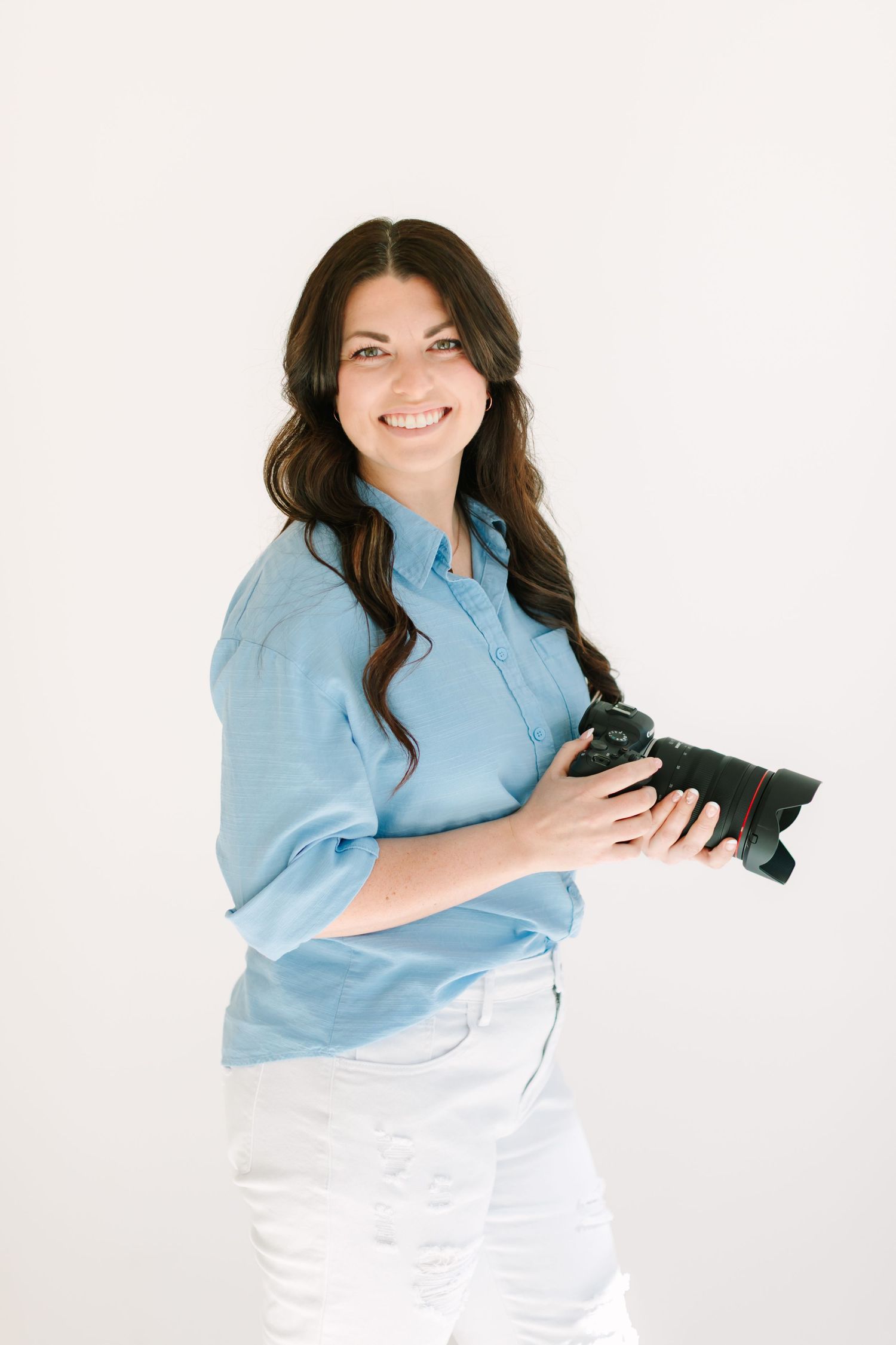 Professional photographer in light blue shirt and white pants holds camera against white background.
