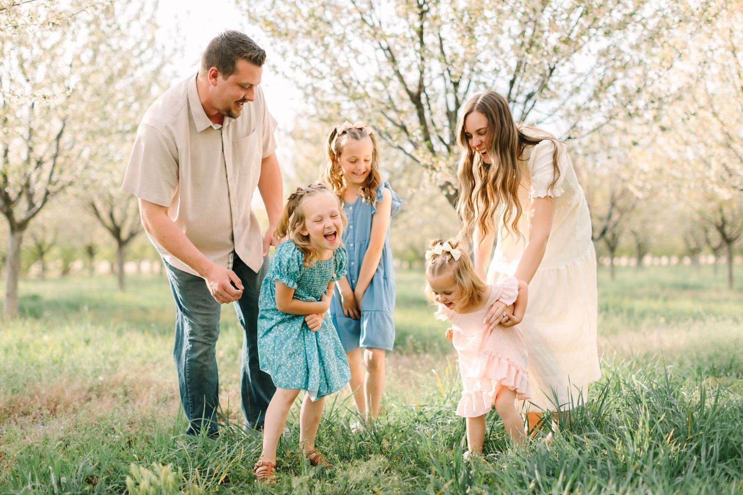 Family of five playing outdoors among blooming spring trees, parents smiling as children laugh and dance in green grass in Utah.