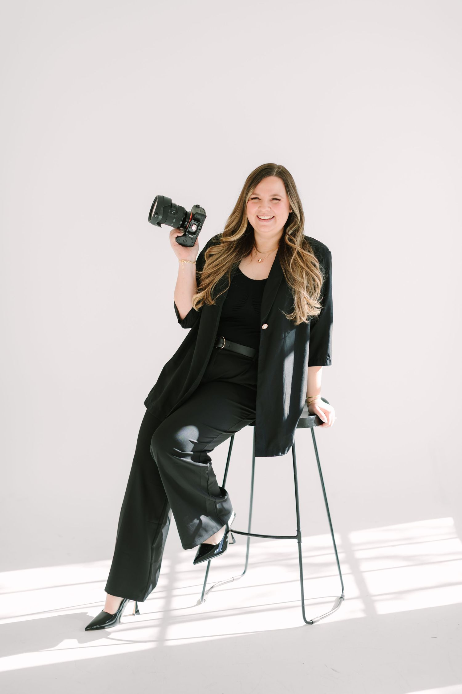Professional photographer in black attire poses with camera while seated on modern chair in white studio setting.