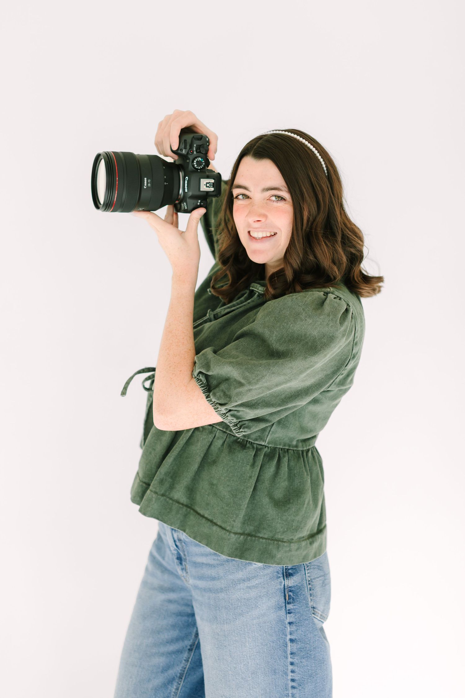 Photographer in olive green sweater and jeans holds professional camera while posing against white background.