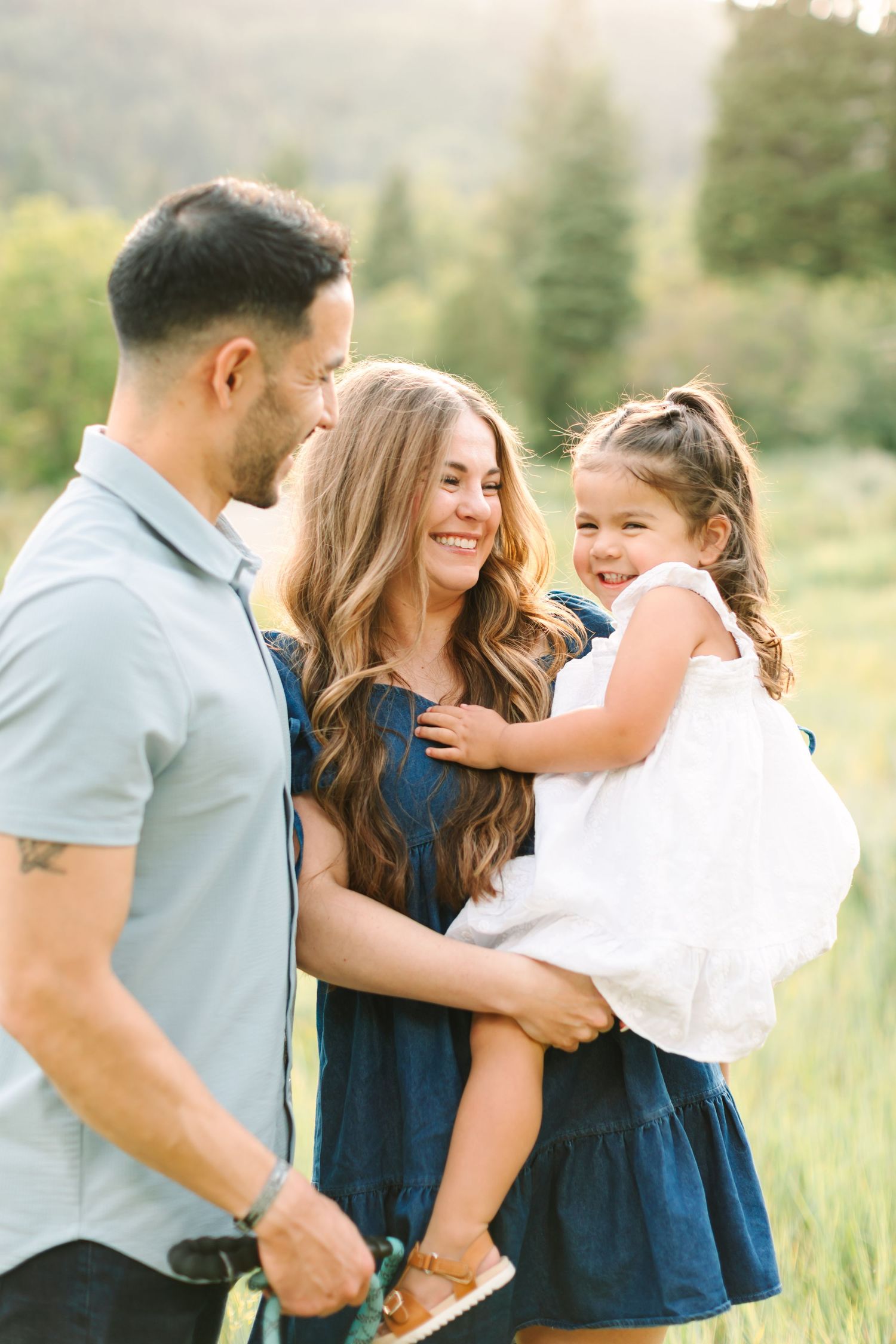 A happy family of three poses outdoors, mom holding toddler daughter in white dress, dad smiling in a sunlit meadow.