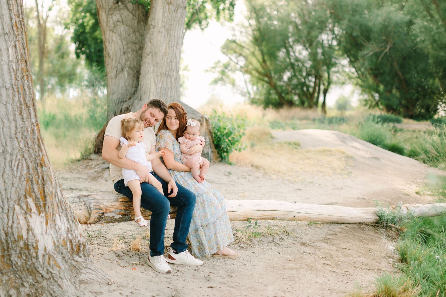 Family of four sitting on a log under a large tree in a natural outdoor setting during a professional photo session.