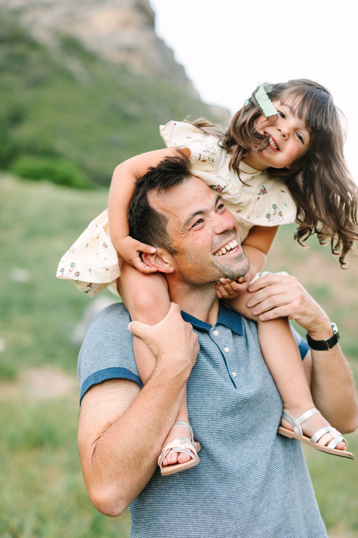A smiling man gives a laughing little girl a piggyback ride outdoors with green hills in the background.