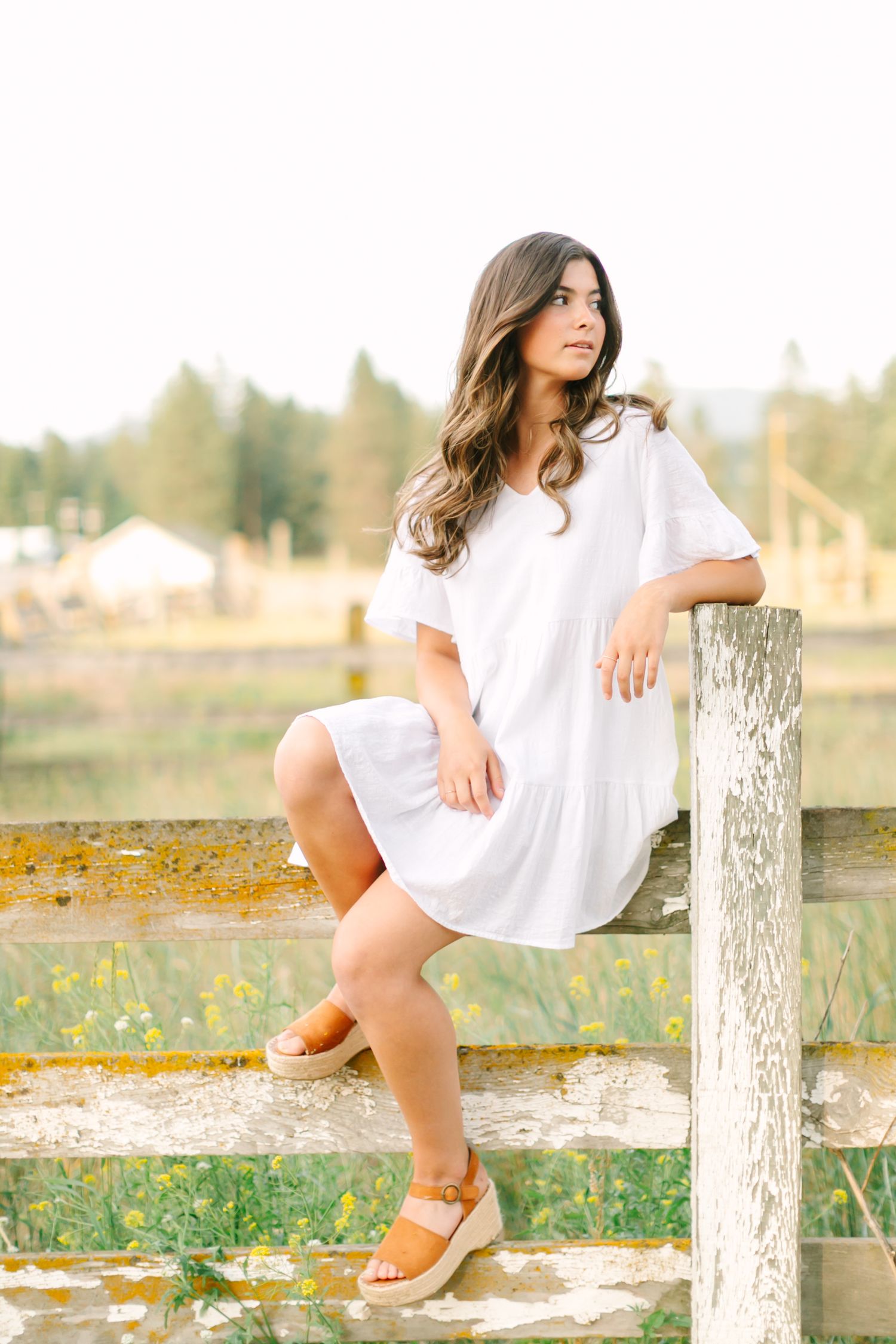 Woman in white dress sits casually on rustic wooden fence surrounded by wildflowers in golden afternoon light.