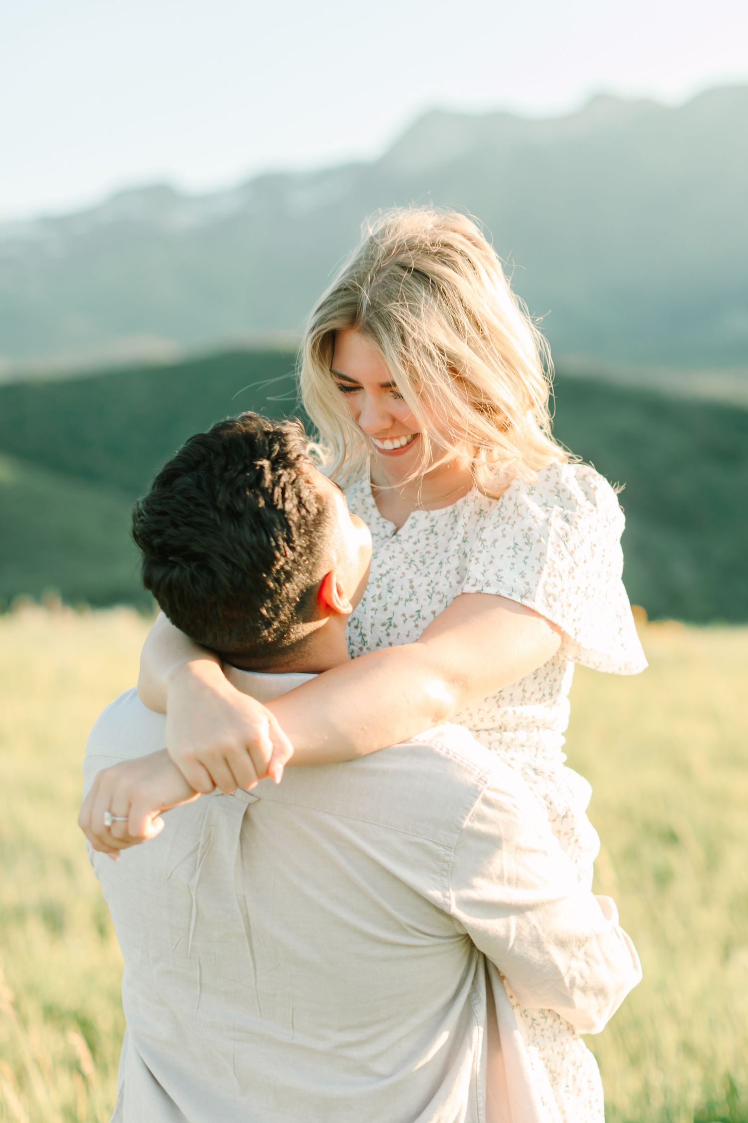 A couple embraces in a scenic mountain meadow at sunset, with golden light illuminating the grassy field.