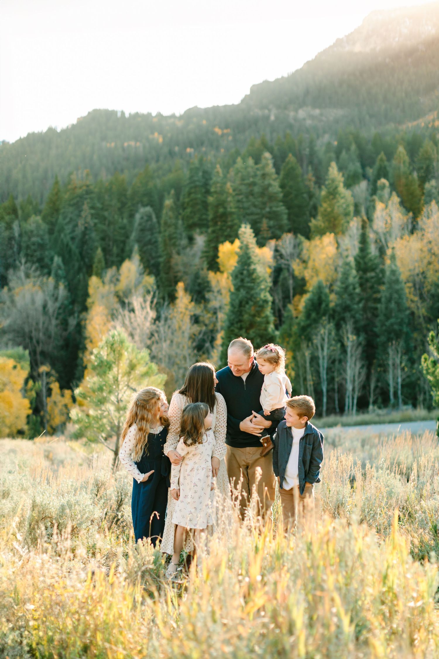 A group stands together in a golden meadow with evergreen forest and mountains in the misty background during sunset.