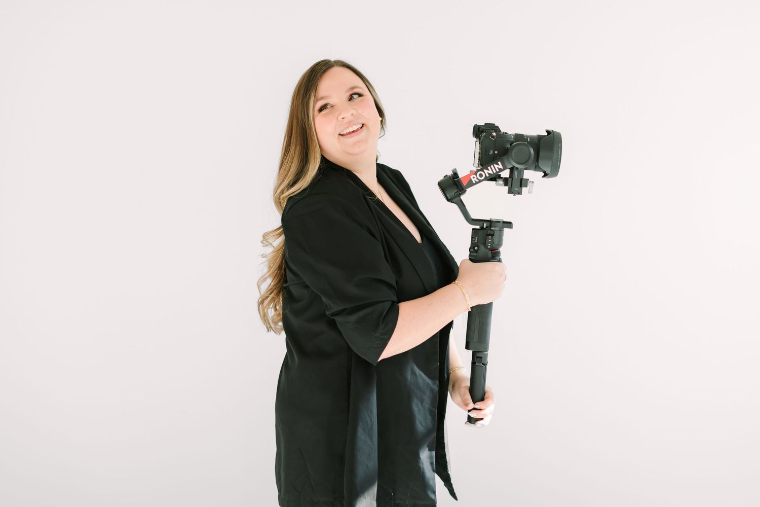 Professional photographer in black blazer holding camera and posing in multiple angles against white background.