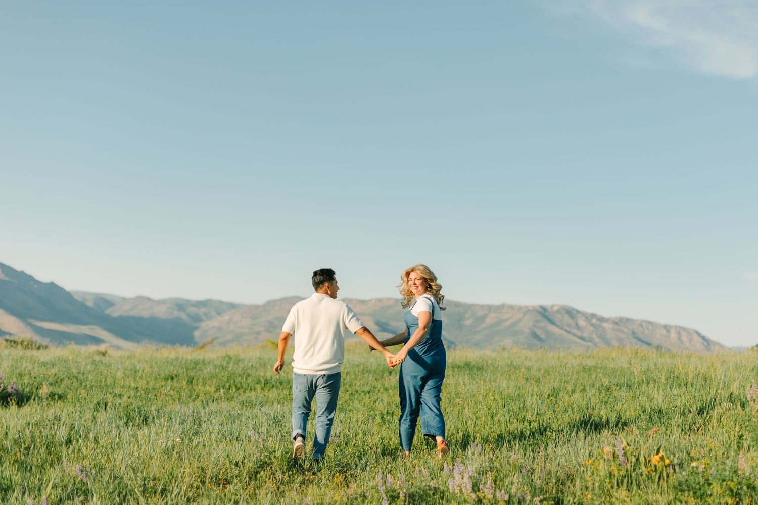 Couple walking hand in hand through a scenic mountain meadow filled with wildflowers on a sunny summer day.
