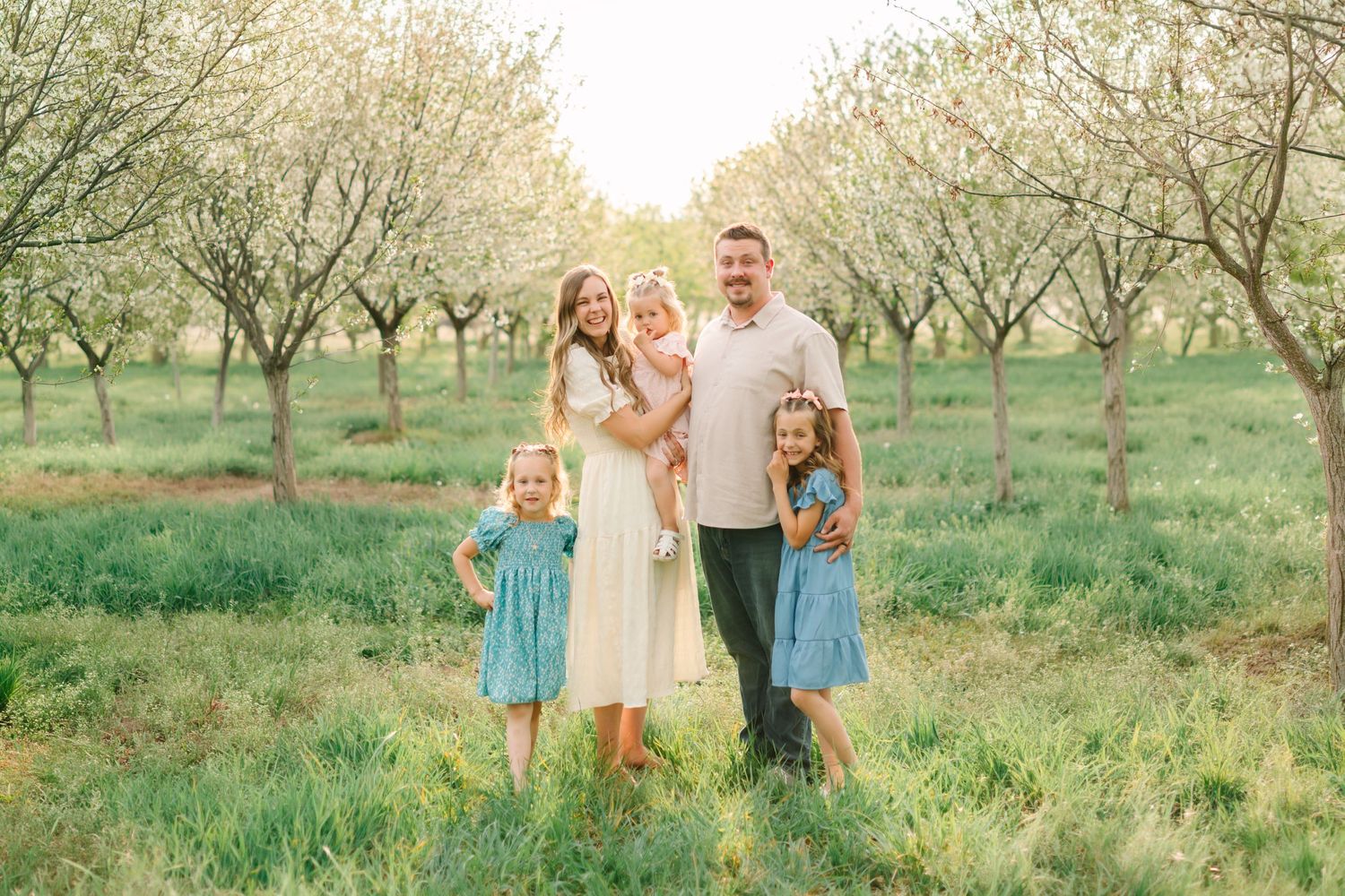 Family of five posing in a sunlit orchard with blooming trees, wearing coordinated outfits in white and blue.