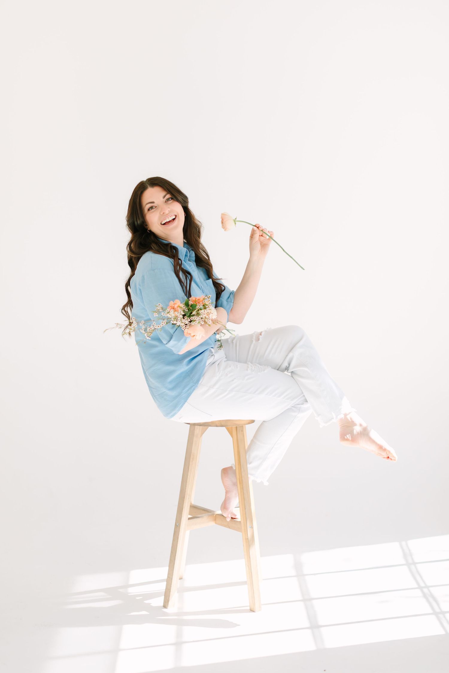 A person in a light blue shirt and white pants sits playfully on a wooden stool against a bright white background.