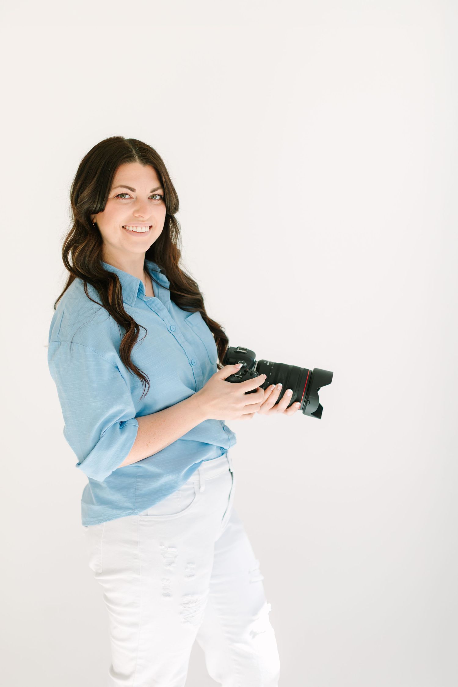 Professional photographer in light blue shirt and white pants poses with camera against white background.