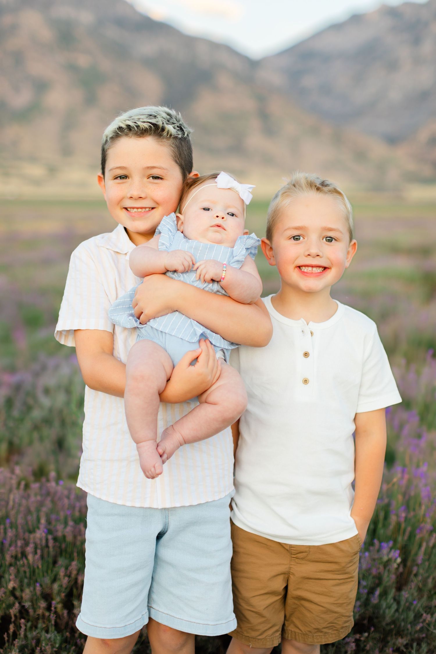 Three young children pose together in a field of purple wildflowers with mountains visible in the background.