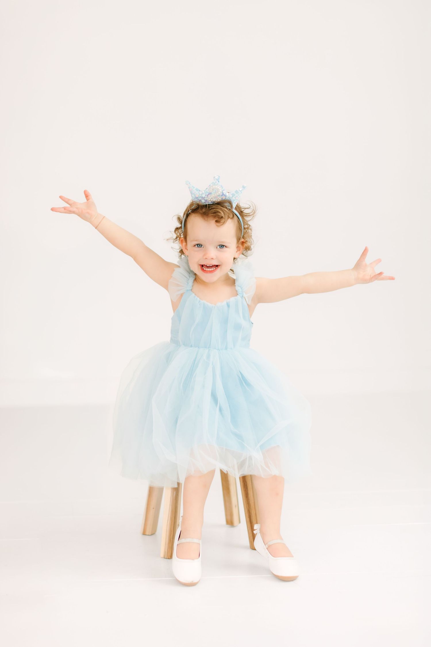 A toddler in a blue tulle dress stands with her arms outstretched, smiling happily for her birthday milestone photoshoot in Utah.