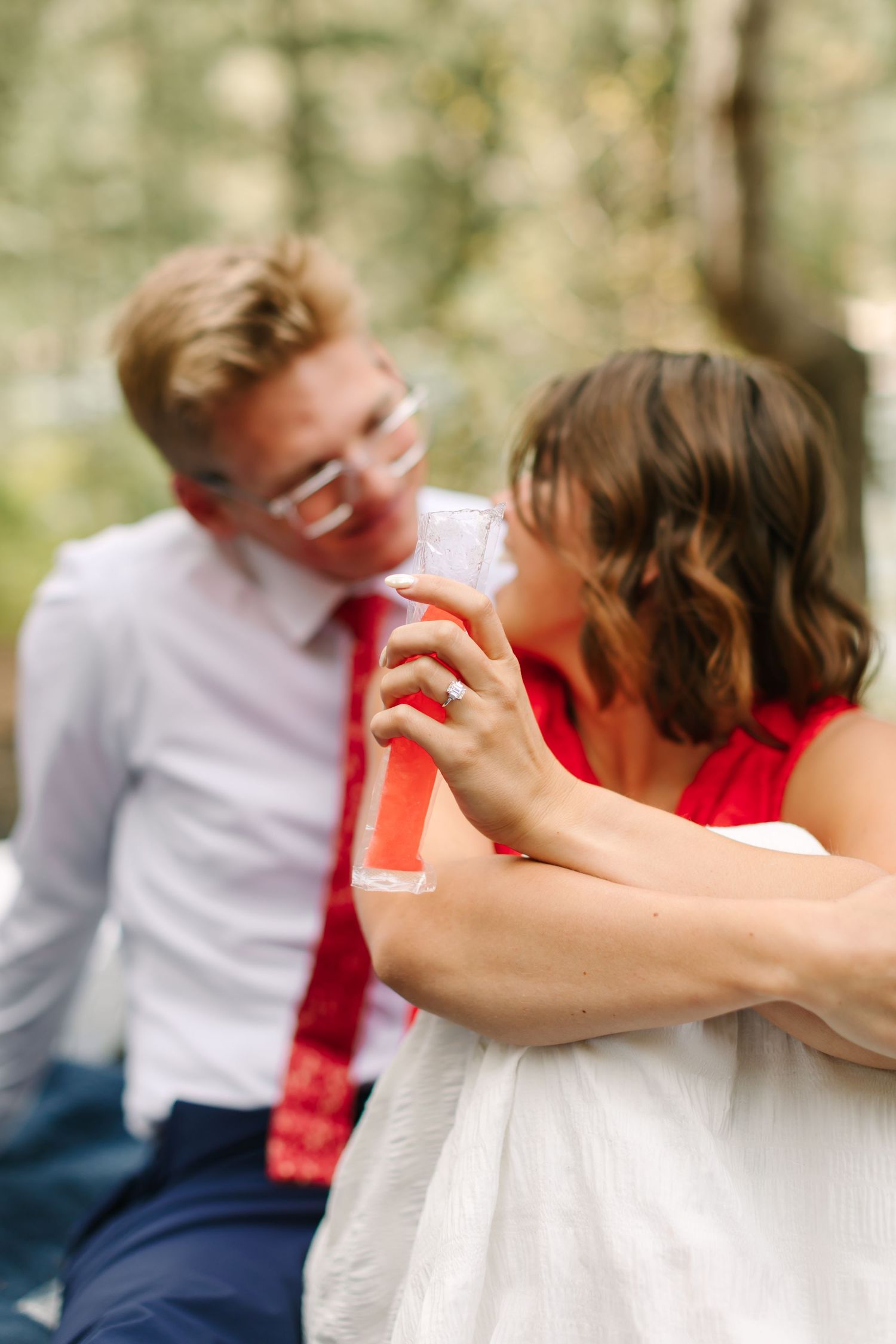 A bride in white dress sips pink drink while groom in red tie smiles softly in outdoor wedding setting.