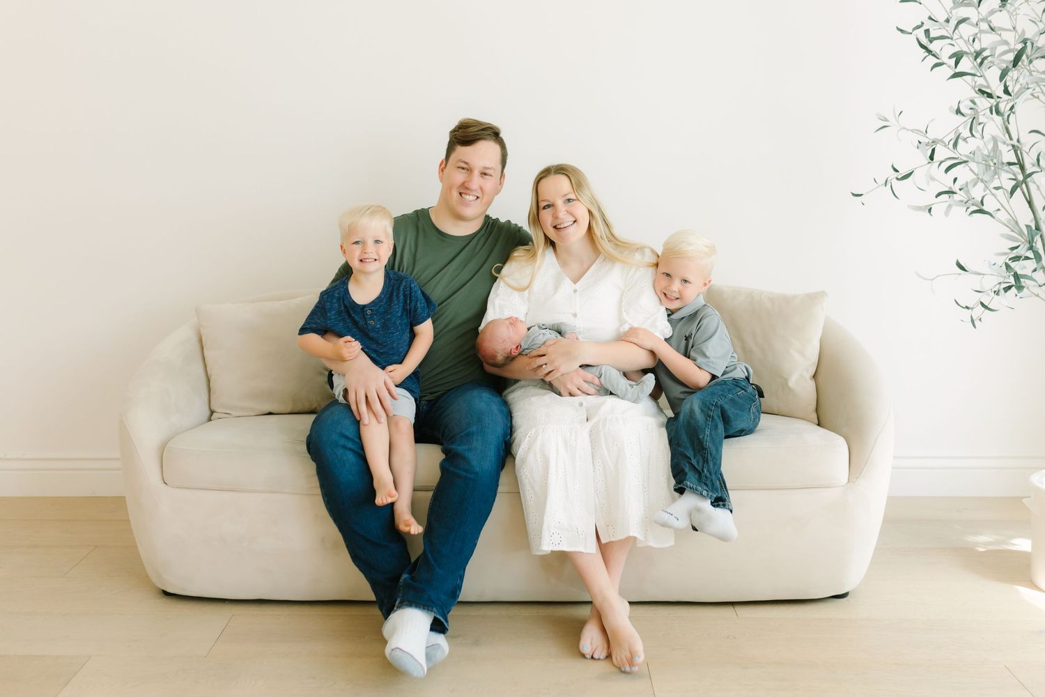 Happy family of five with newborn baby posing together on a beige sofa in a bright, minimalist studio setting.