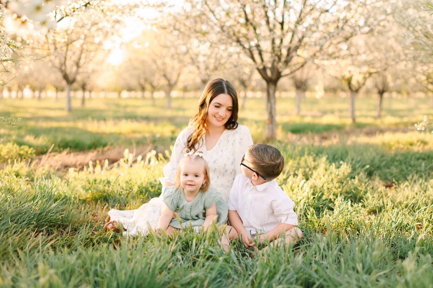 A young family sits together in a sunlit orchard with blooming trees, mom in white dress with toddler and child on green grass.