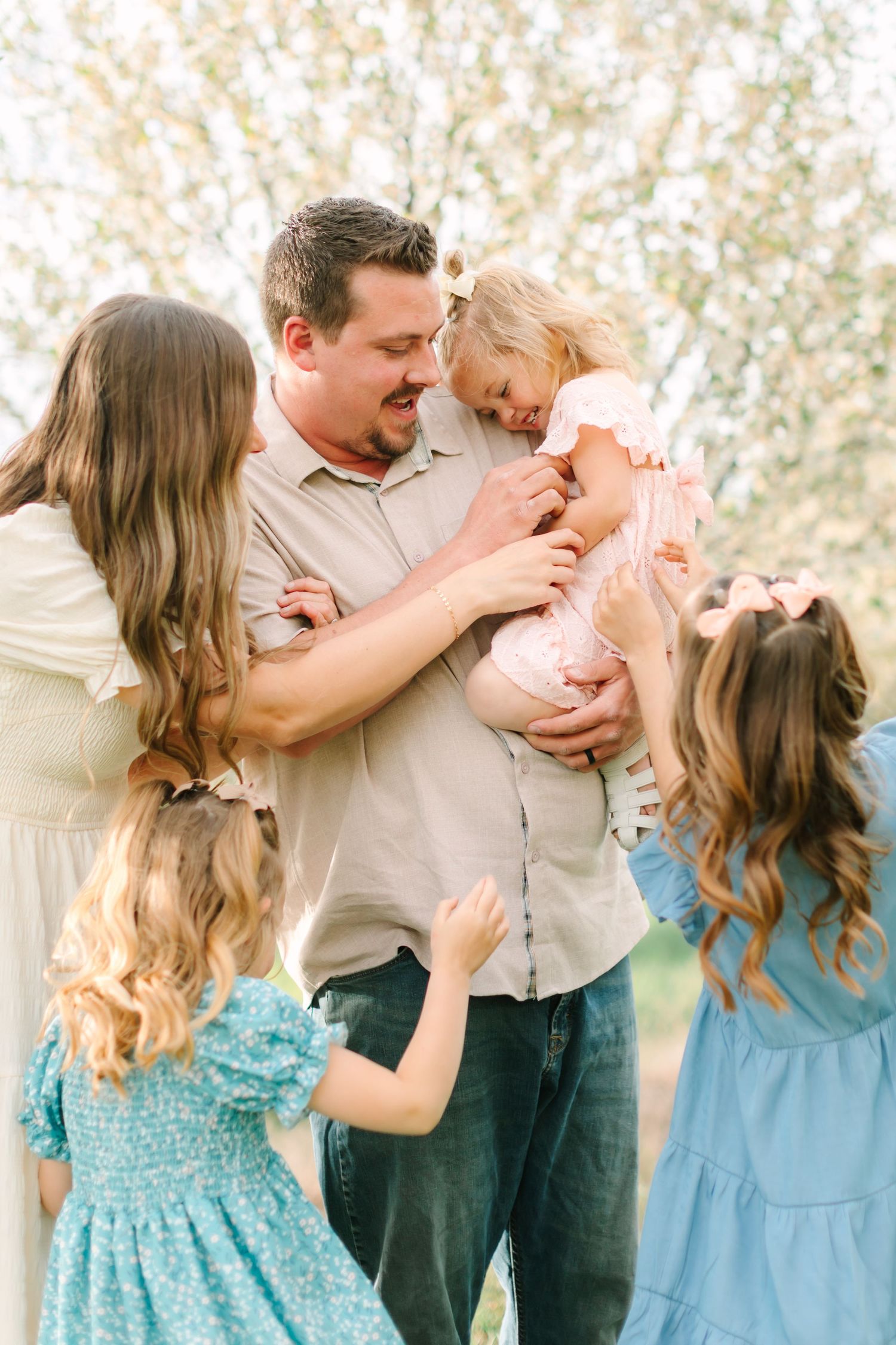 A happy family with a father holding a toddler girl surrounded by his wife and daughters outdoors.
