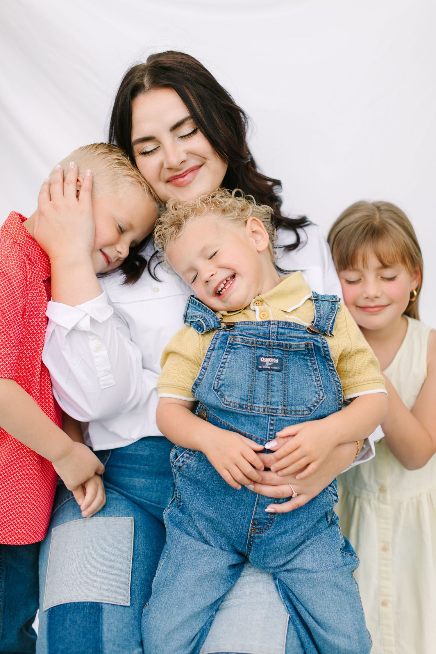 A smiling brunette mother hugs three happy children in a bright white studio family portrait.