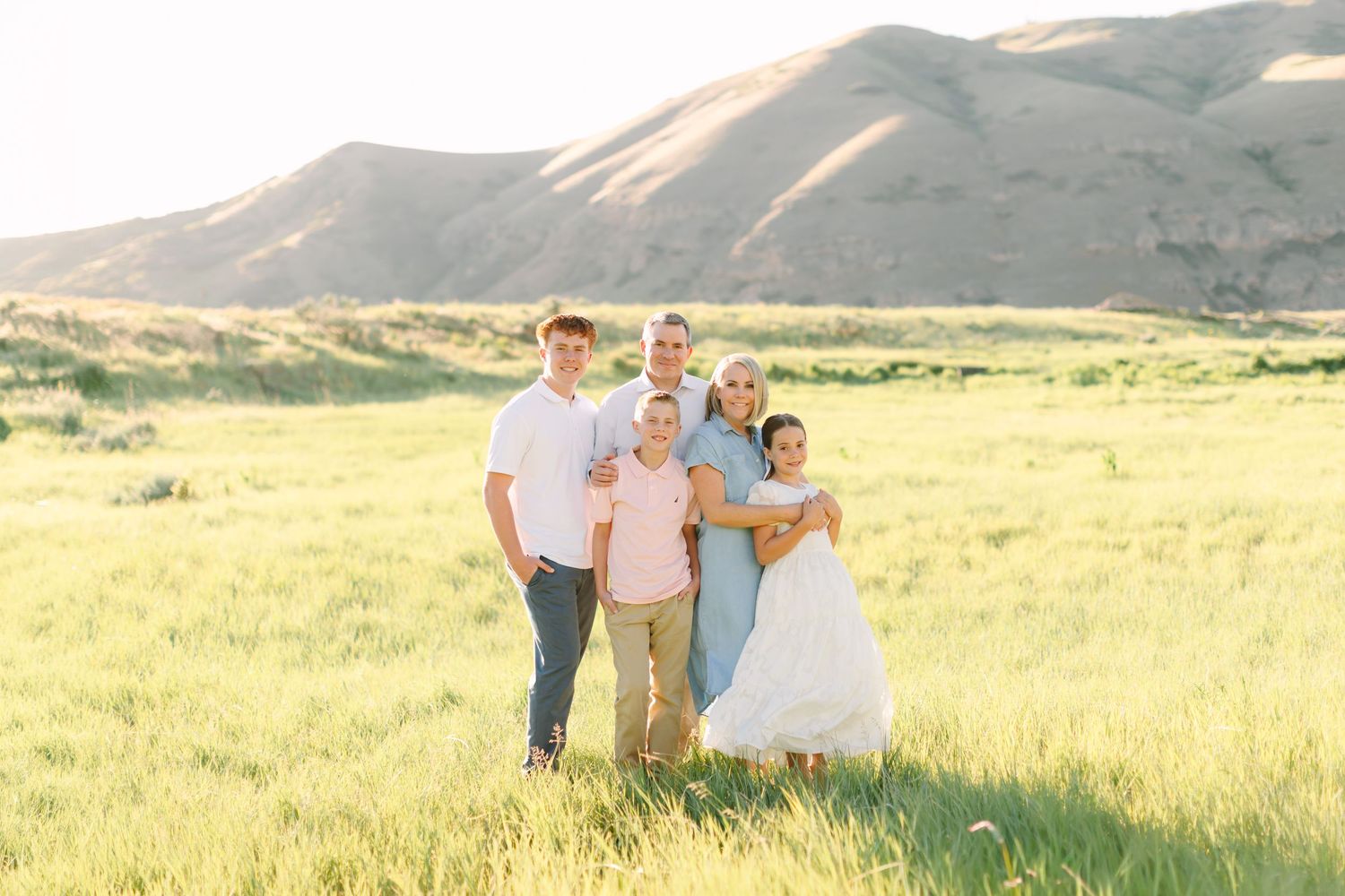 Family of five posing together in a lush green field with scenic mountain backdrop in warm golden sunlight.