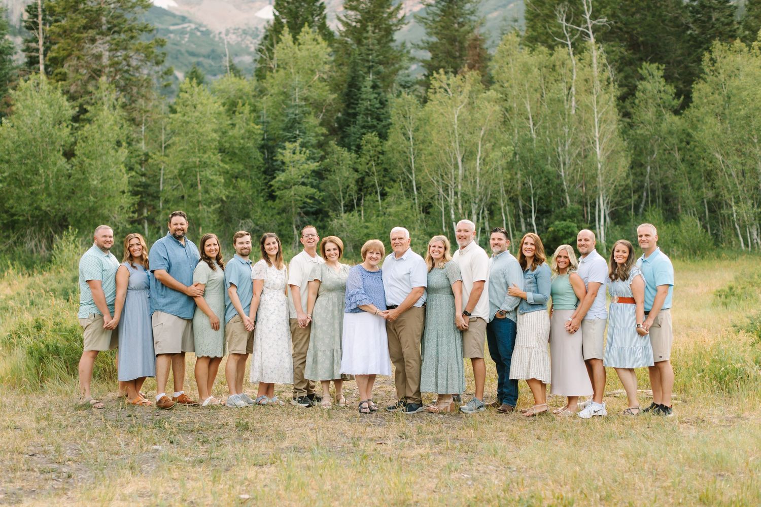 Large family group smiling outdoors in coordinated sage green and white outfits with lush green trees behind in Provo, Utah.