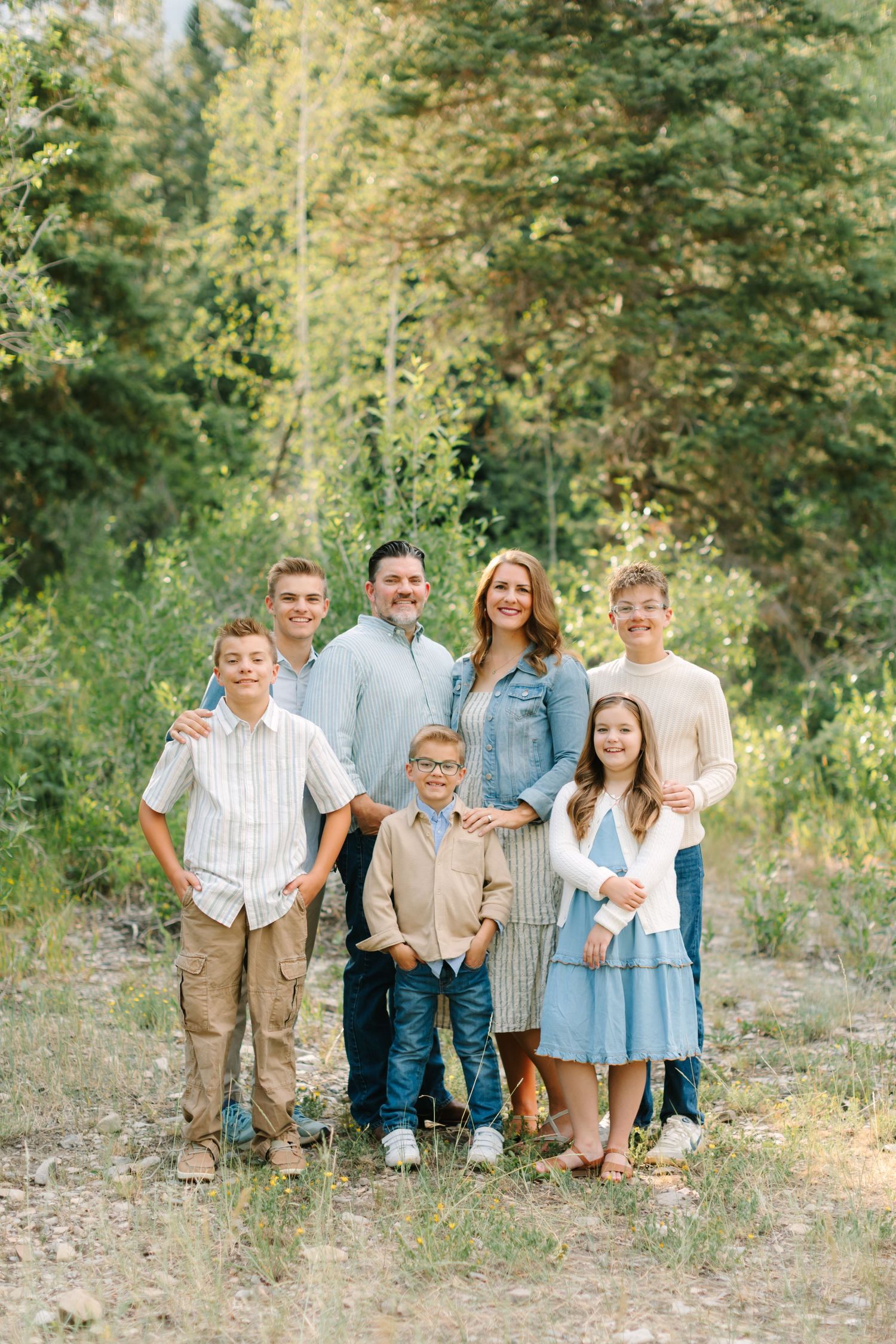 Family of seven posing outdoors in coordinated neutral outfits surrounded by lush green trees in a natural setting.