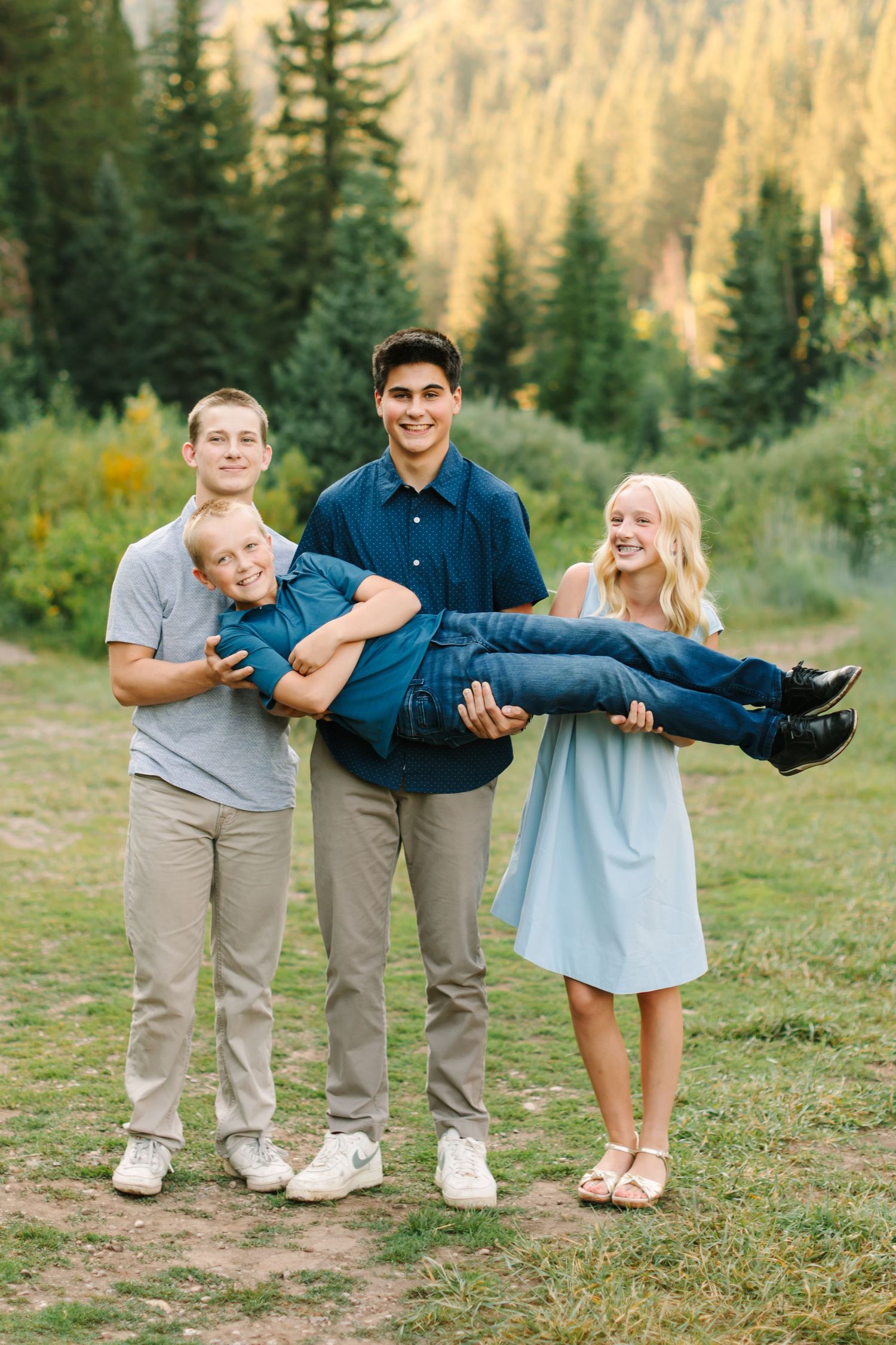 Four siblings pose outdoors, two teens holding a younger boy, with pine trees and golden foliage in the background.