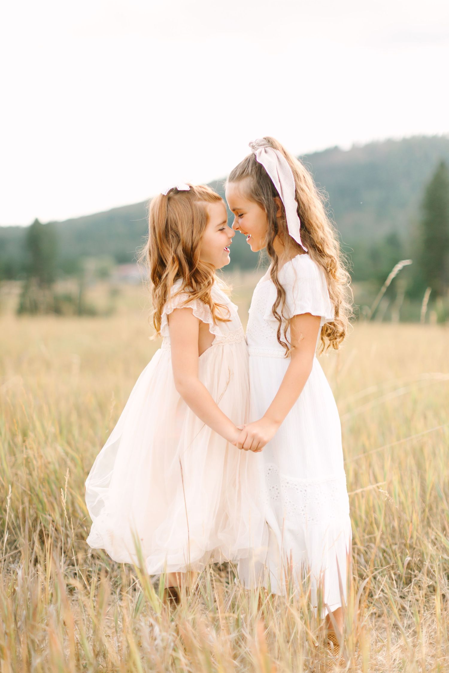 Two young women in white dresses share a tender moment in a sunlit meadow.