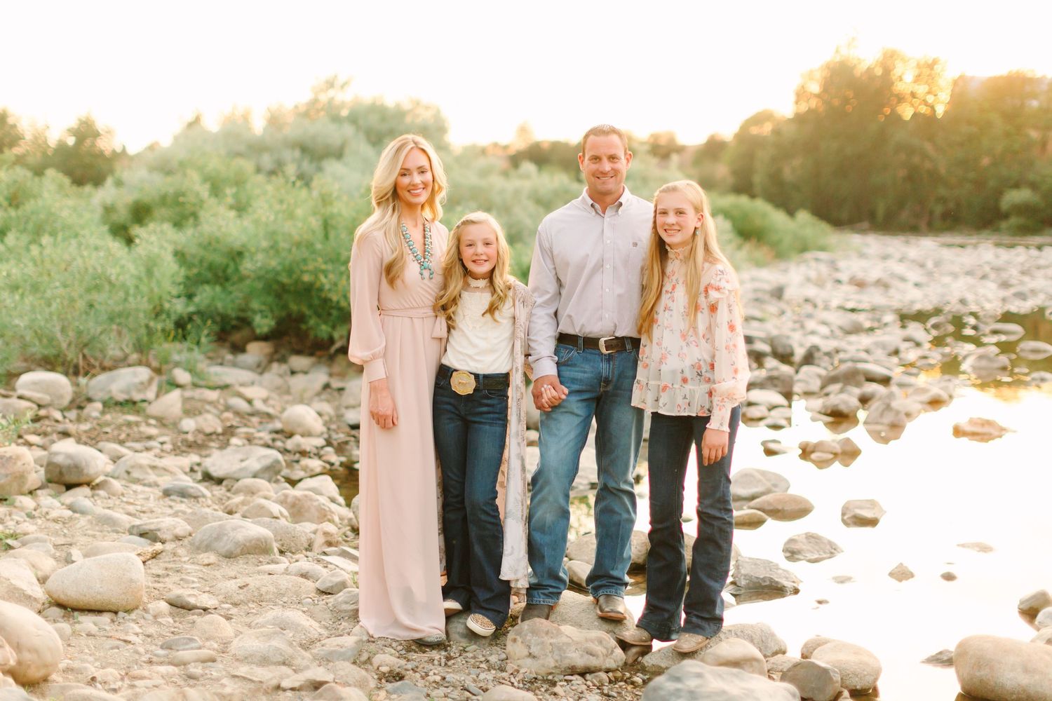 Family wearing coordinated neutral outfits poses together on rocky beach during golden hour sunset.
