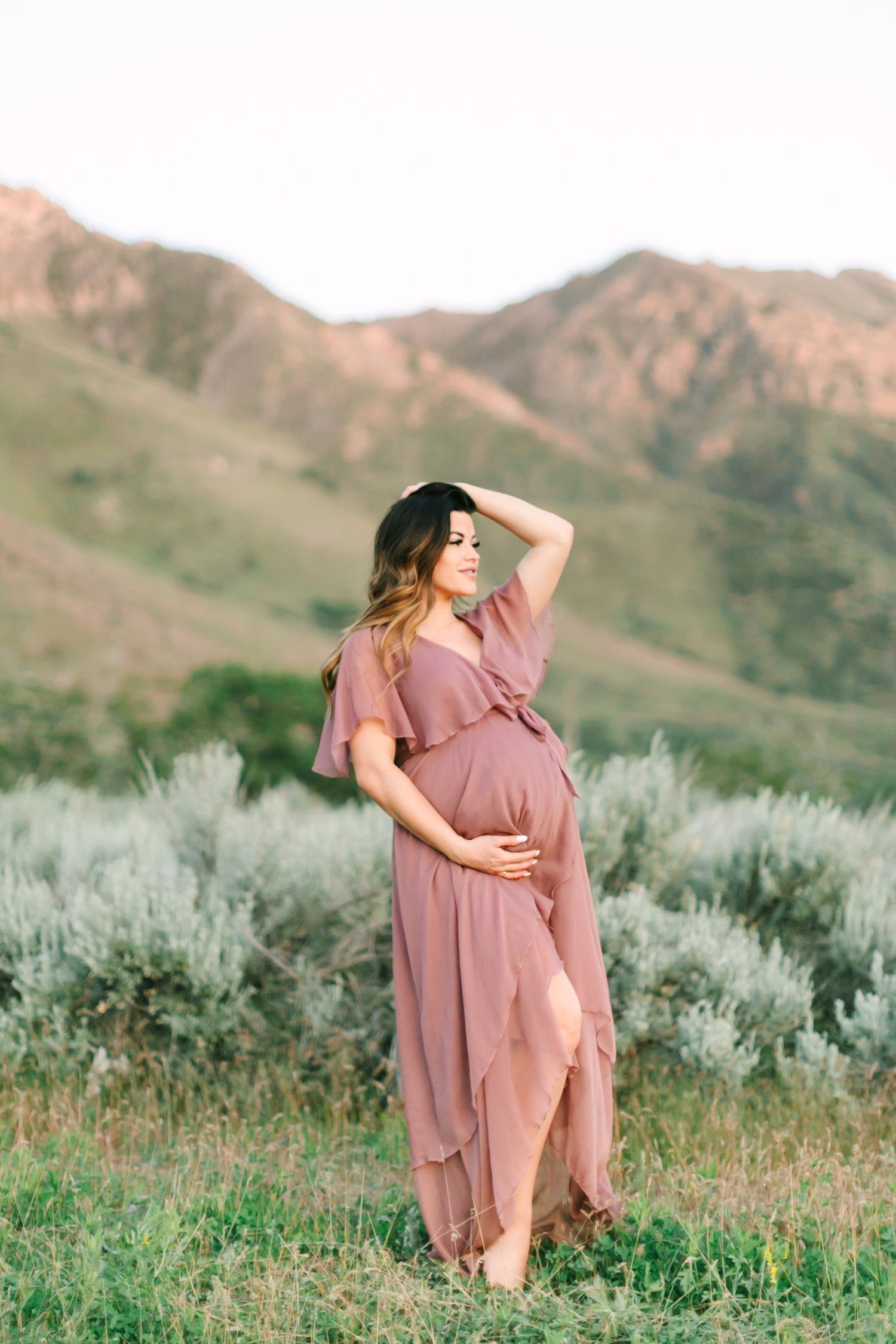 A person in a mauve dress poses gracefully against a backdrop of sage brush and rolling desert hills at sunset.