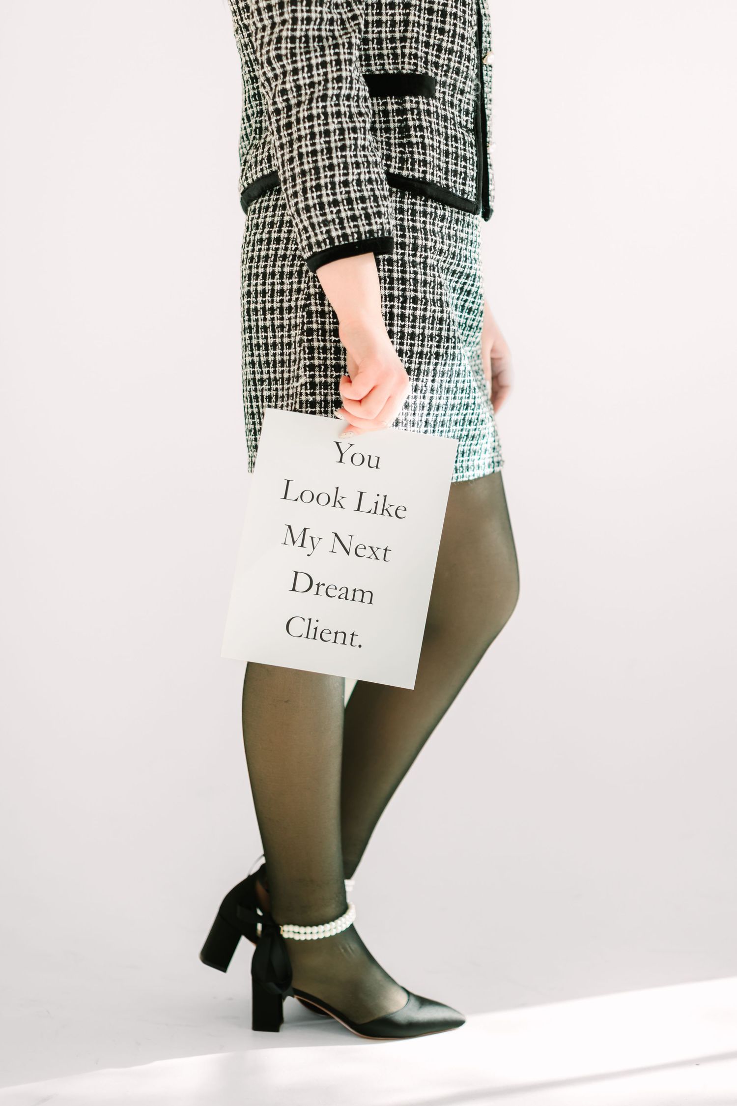 Person wearing plaid jacket and tights holds sign saying 'You Look Like My Next Dream Client' with pearl-adorned heels.