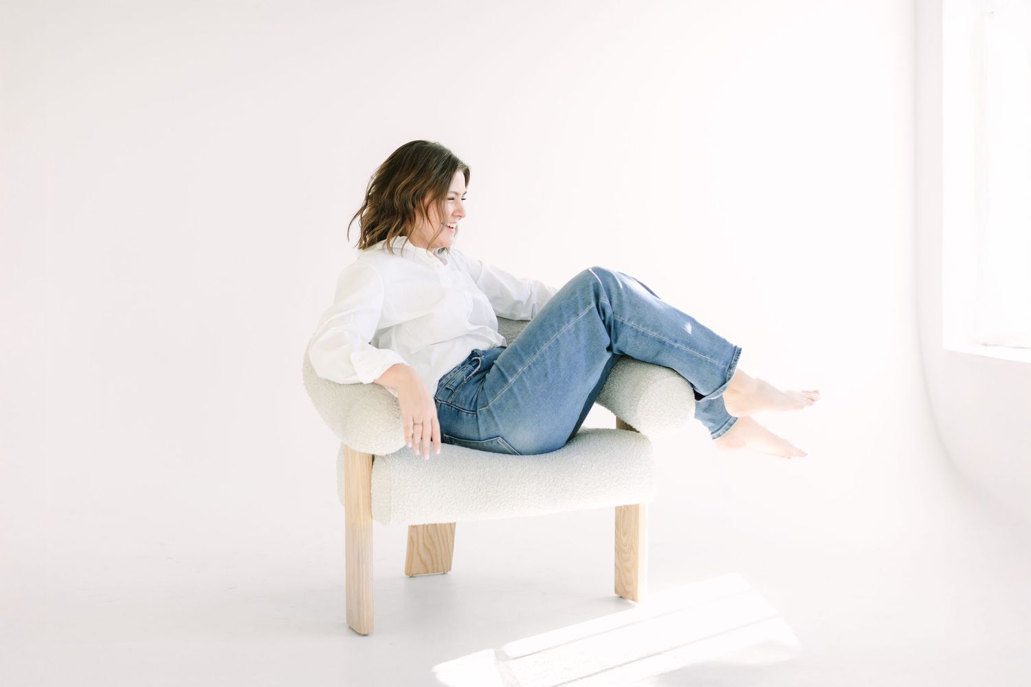 Person in casual white sweater and jeans relaxes on minimalist white bench in bright studio setting.