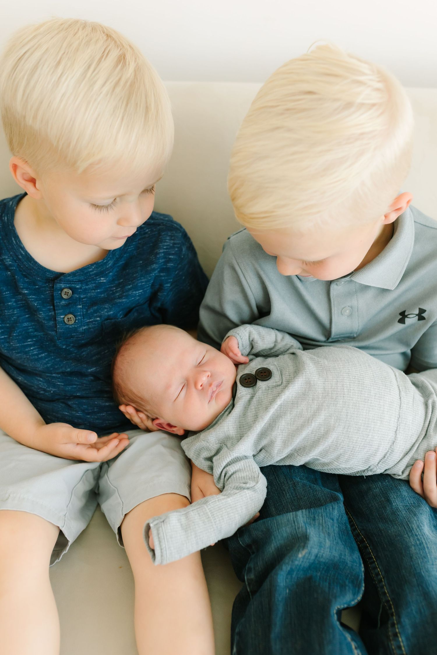 Two blonde toddler boys gently hold their sleeping newborn sibling in gray onesie during a family portrait session.
