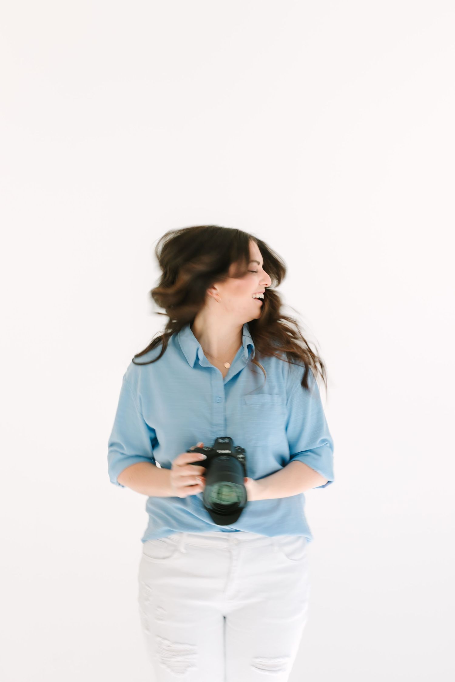 Someone in casual blue shirt and white jeans holding camera while posing against white backdrop.