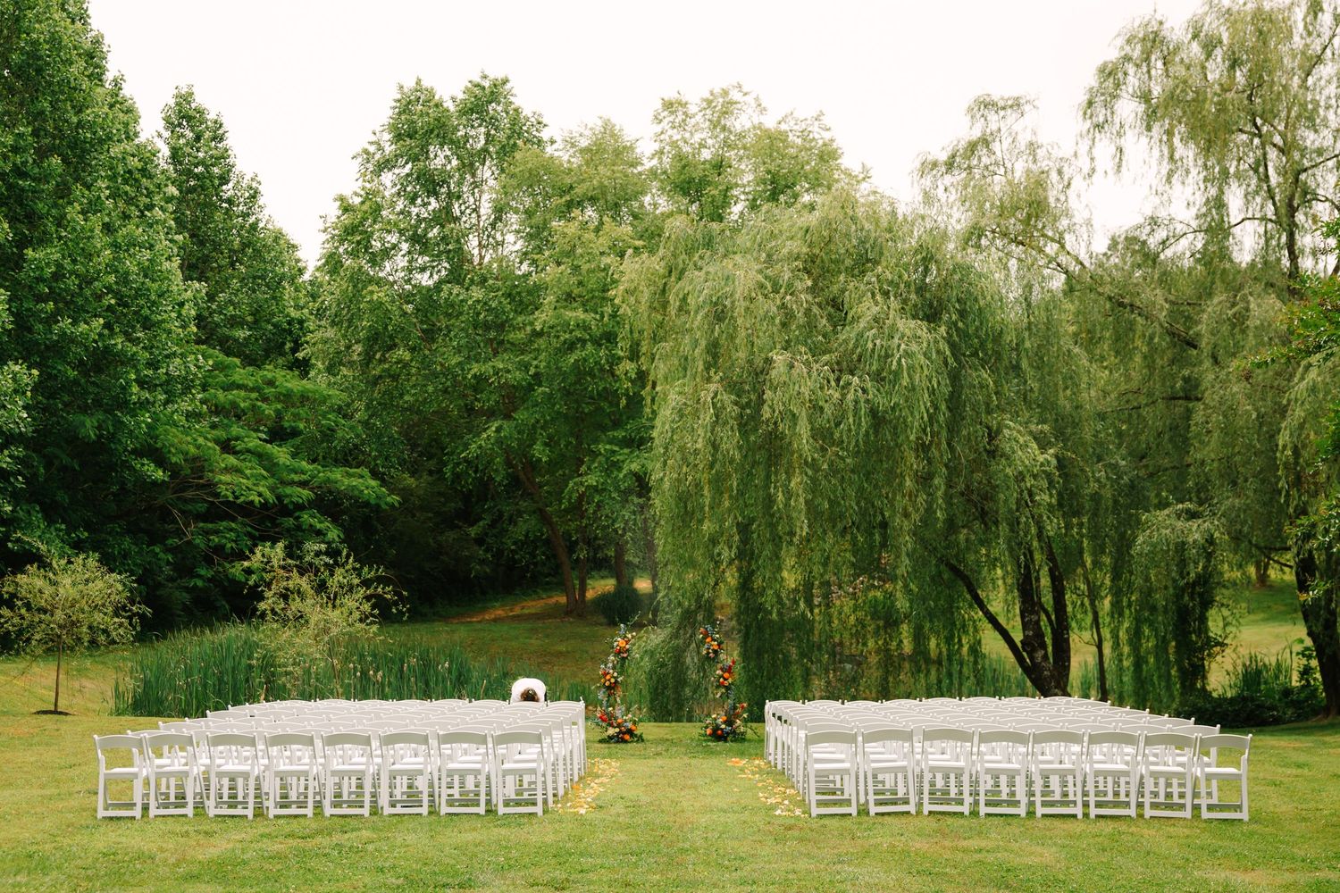 Outdoor wedding ceremony setup with white chairs arranged in rows under weeping willow trees in a lush green garden.